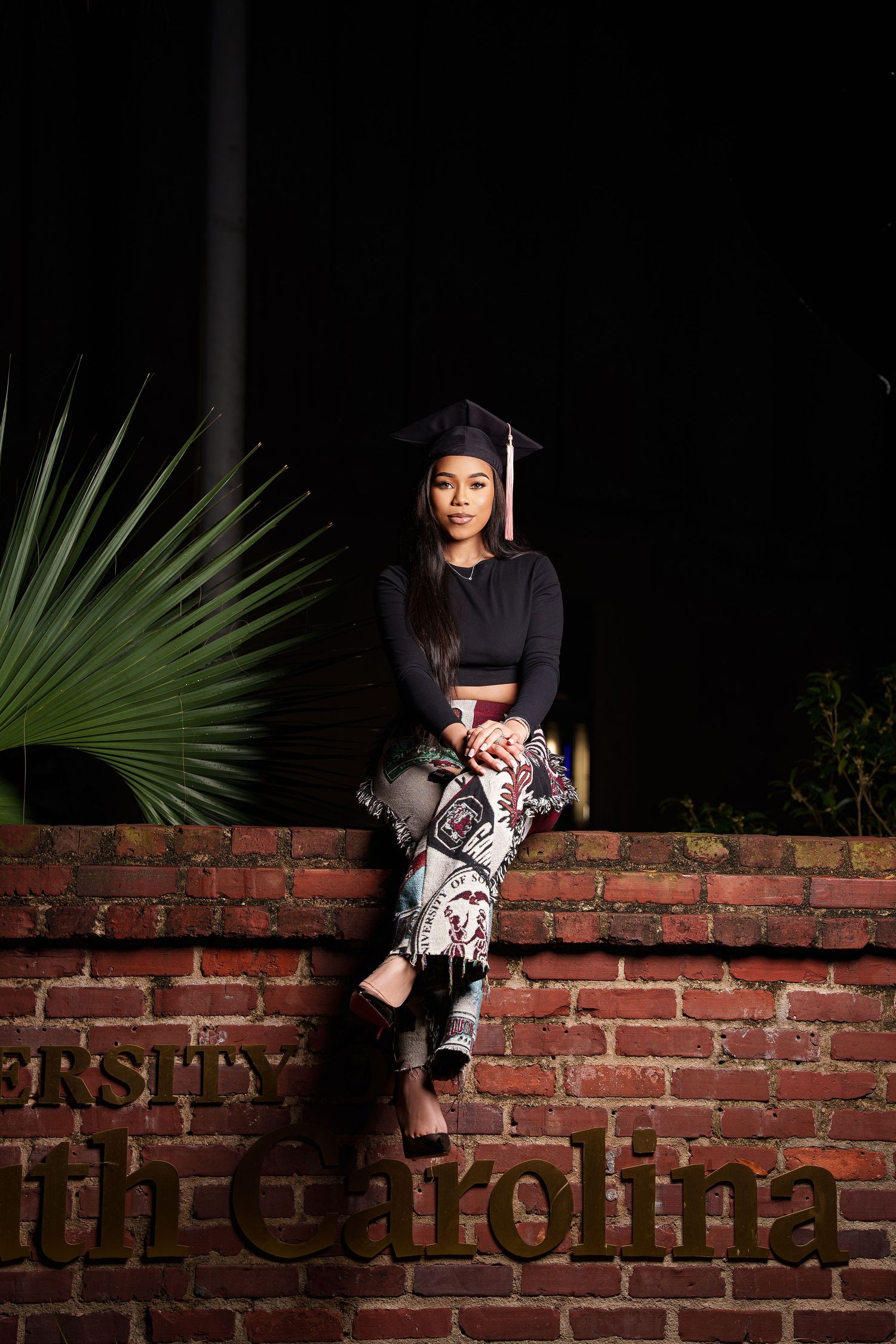 A graduate sits on a brick wall at night with palm fronds visible in the background.