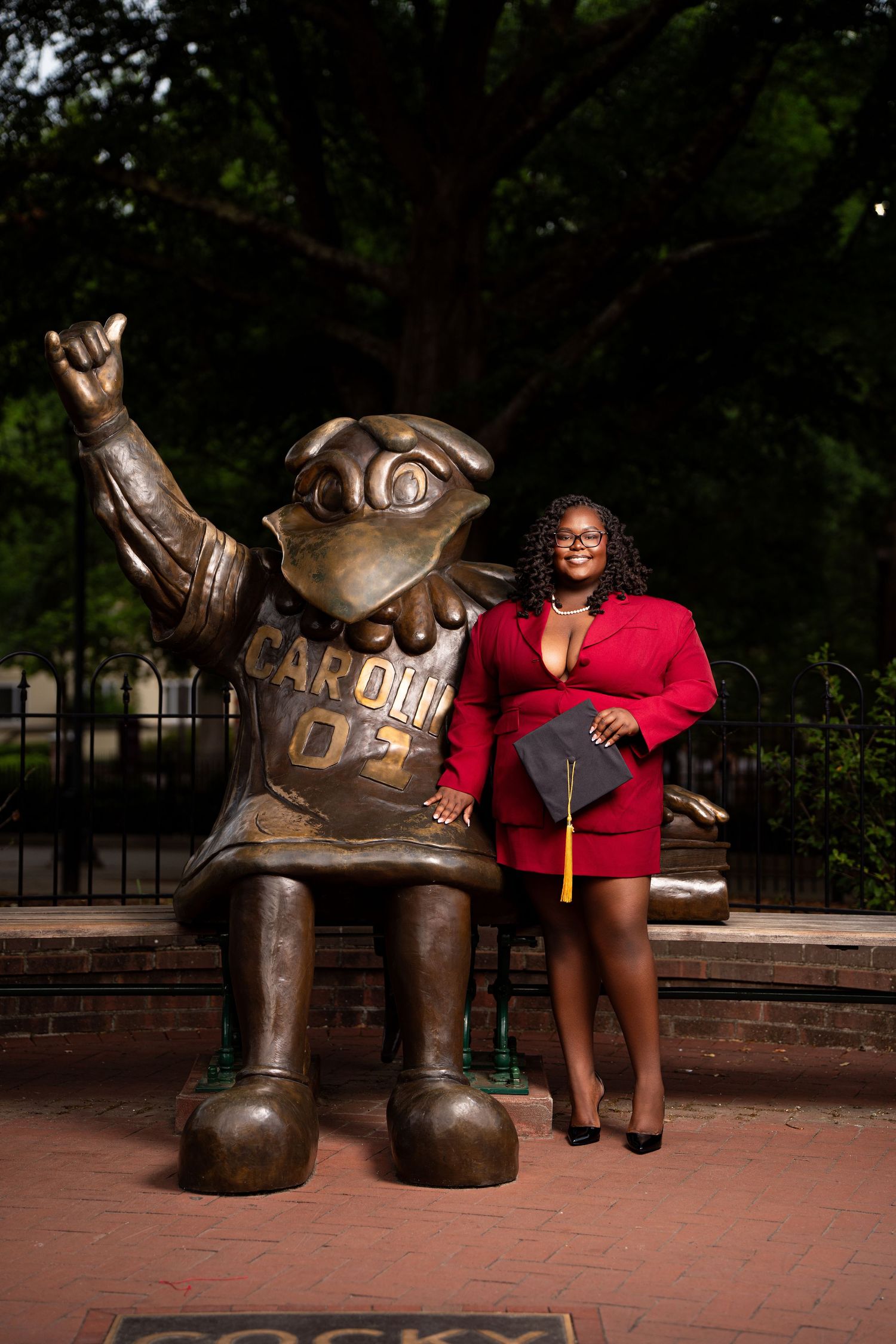 Graduate in red dress holds cap beside University of South Carolina Cocky mascot bronze statue on campus.