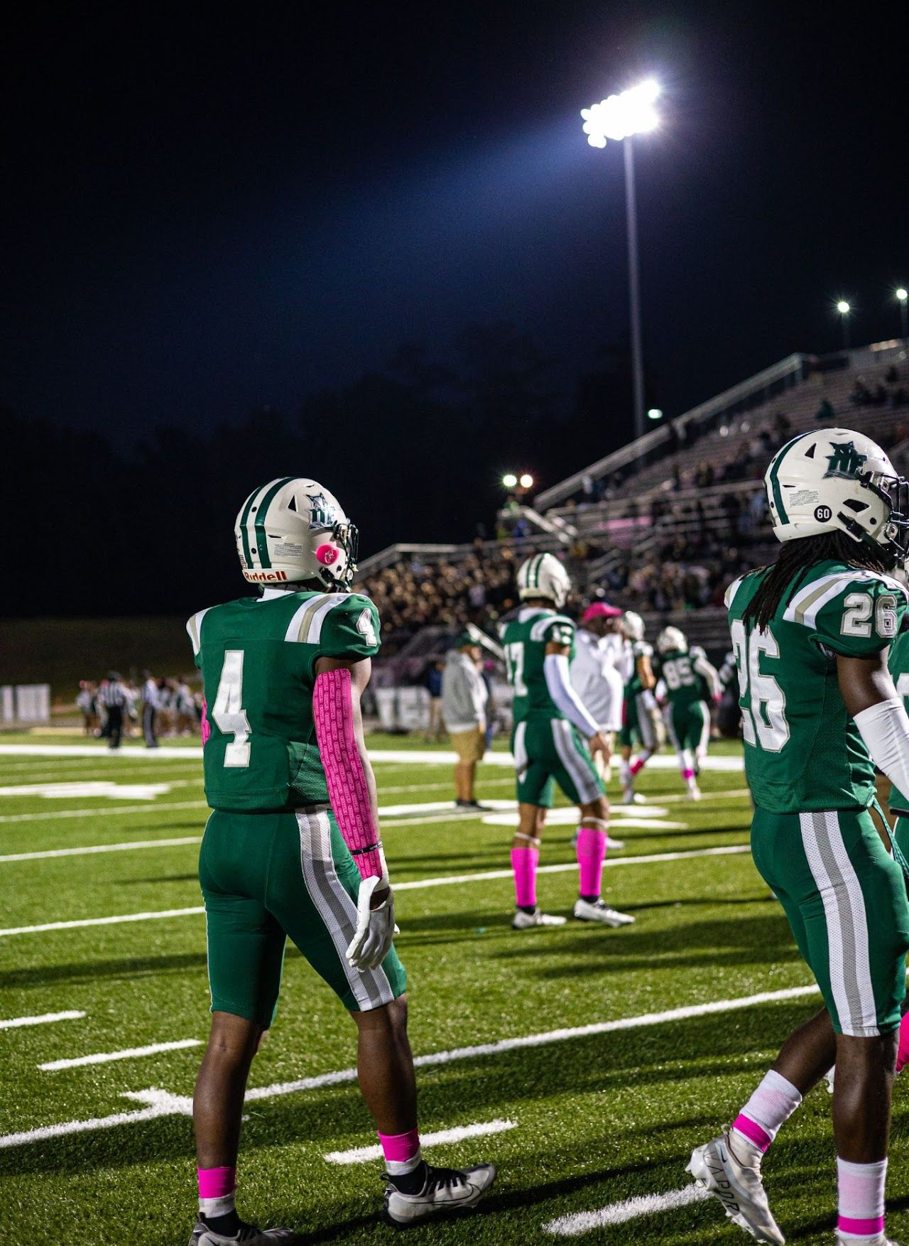 Football players on a night field wearing green uniforms with pink socks during a game under stadium lights.  DFHS