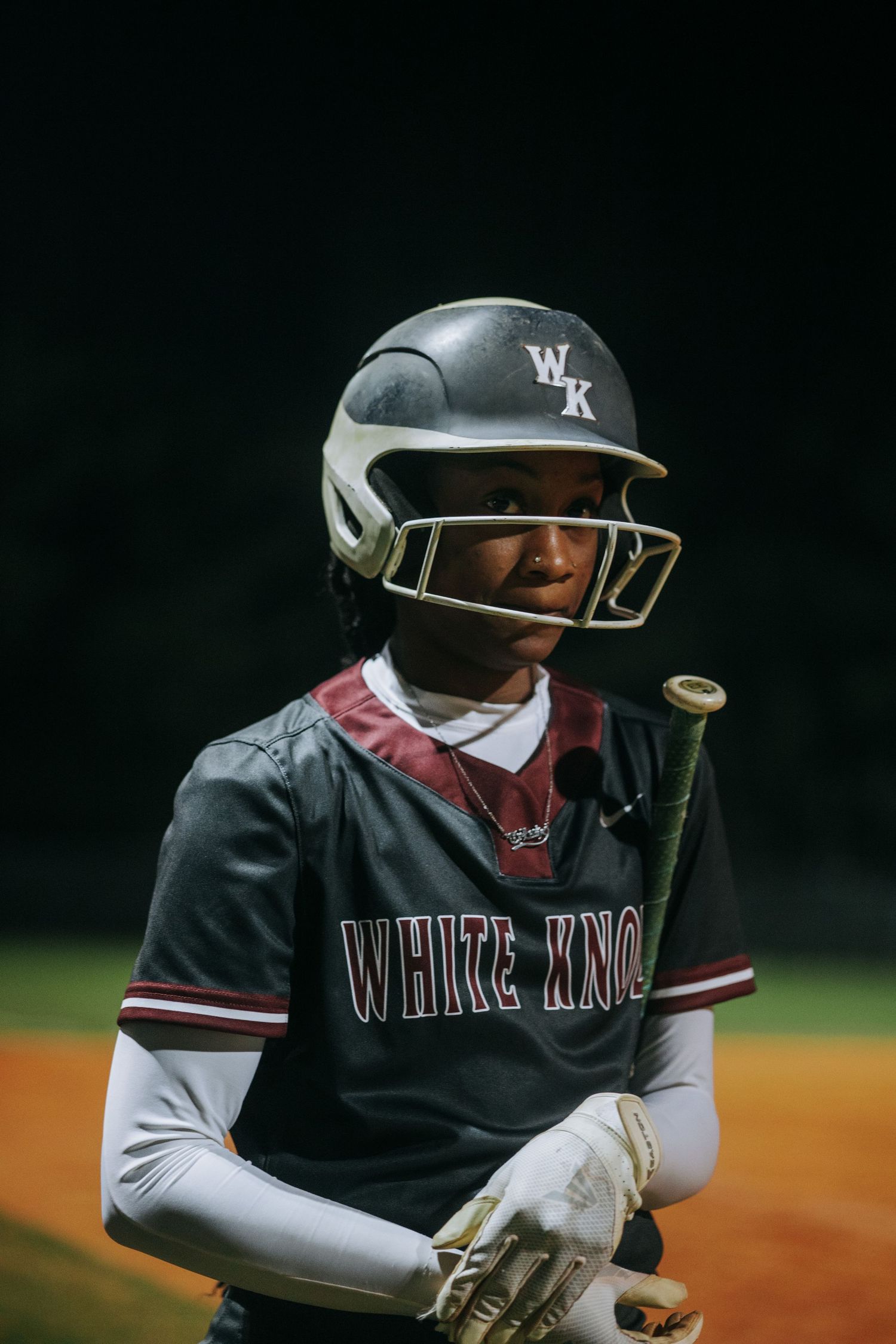Baseball player in White Knot uniform and helmet prepares to bat during nighttime game.