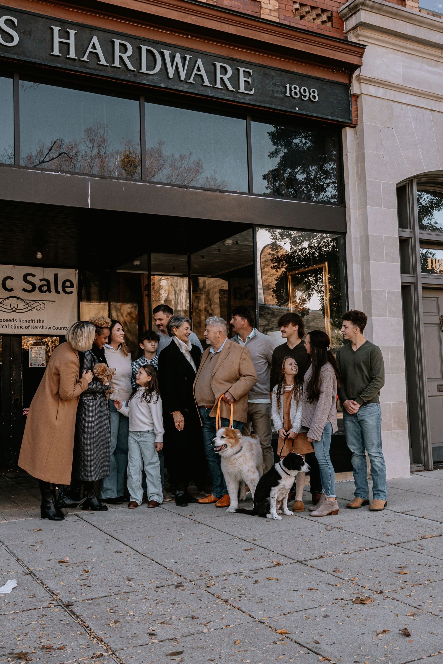 A line of people waiting outside a hardware store storefront on a sidewalk with fallen autumn leaves.