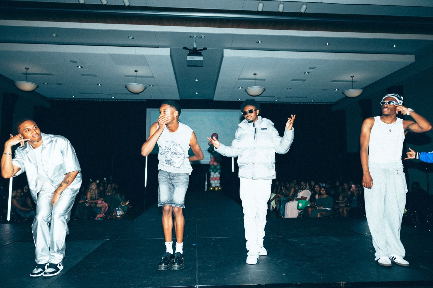 Four performers in white outfits dancing on stage during a live hip-hop performance in a dimly lit venue.