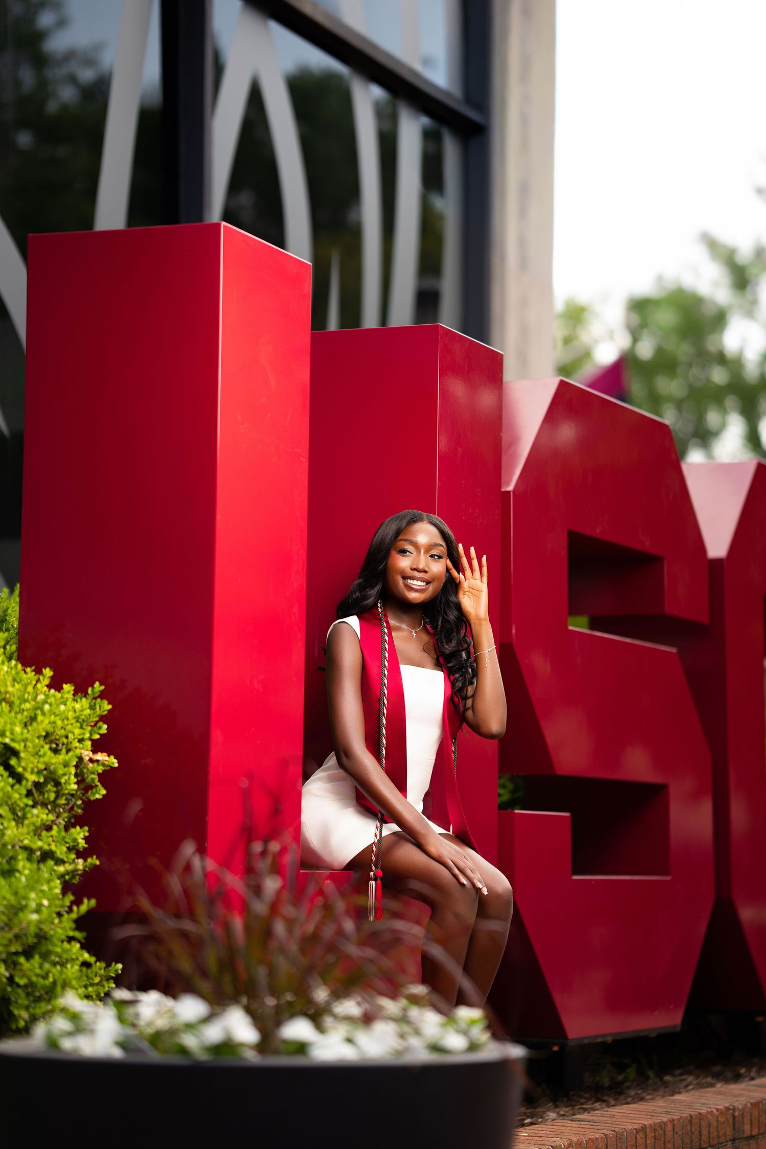 A smiling graduate in a white dress with red tassel sits beside large red letters outdoors.
