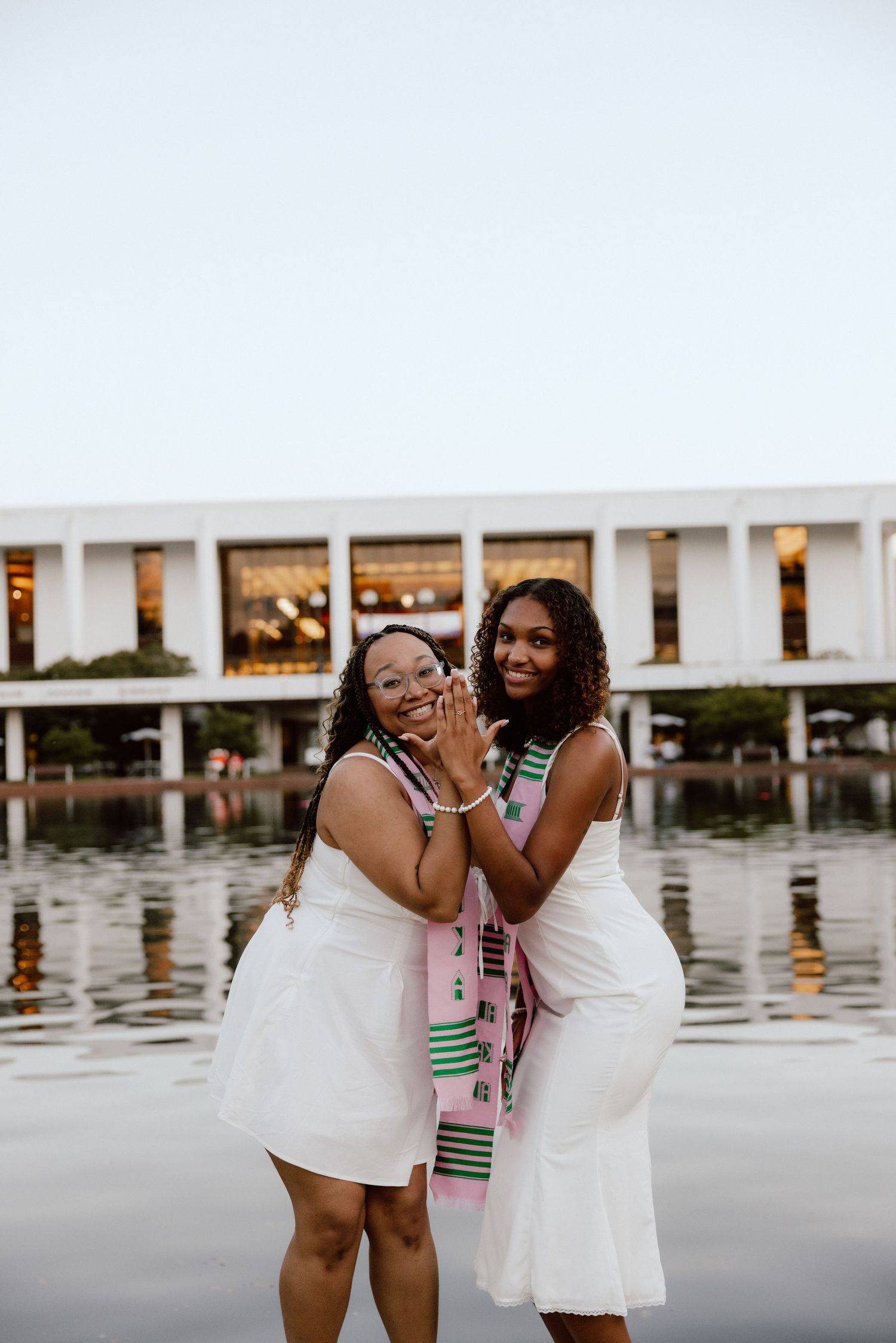 Two young women in white dresses and kente stoles celebrate graduation near a reflective pool.