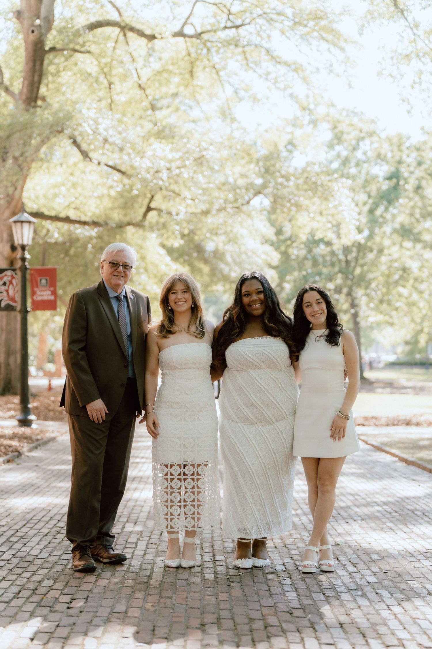 A man in a brown suit and three women in white dresses pose together on a brick path surrounded by trees.