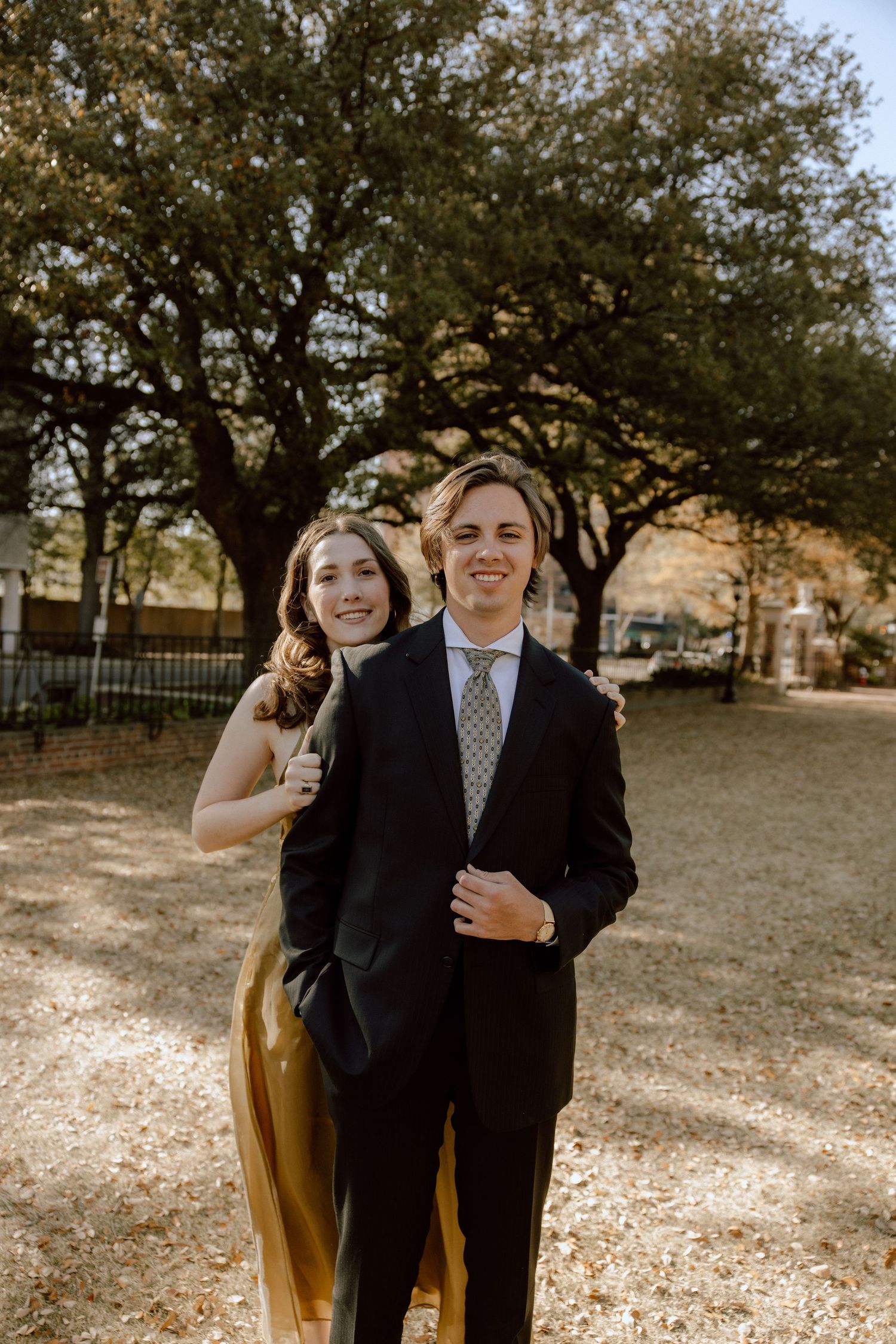 A couple smiles together outdoors under trees in formal wear.