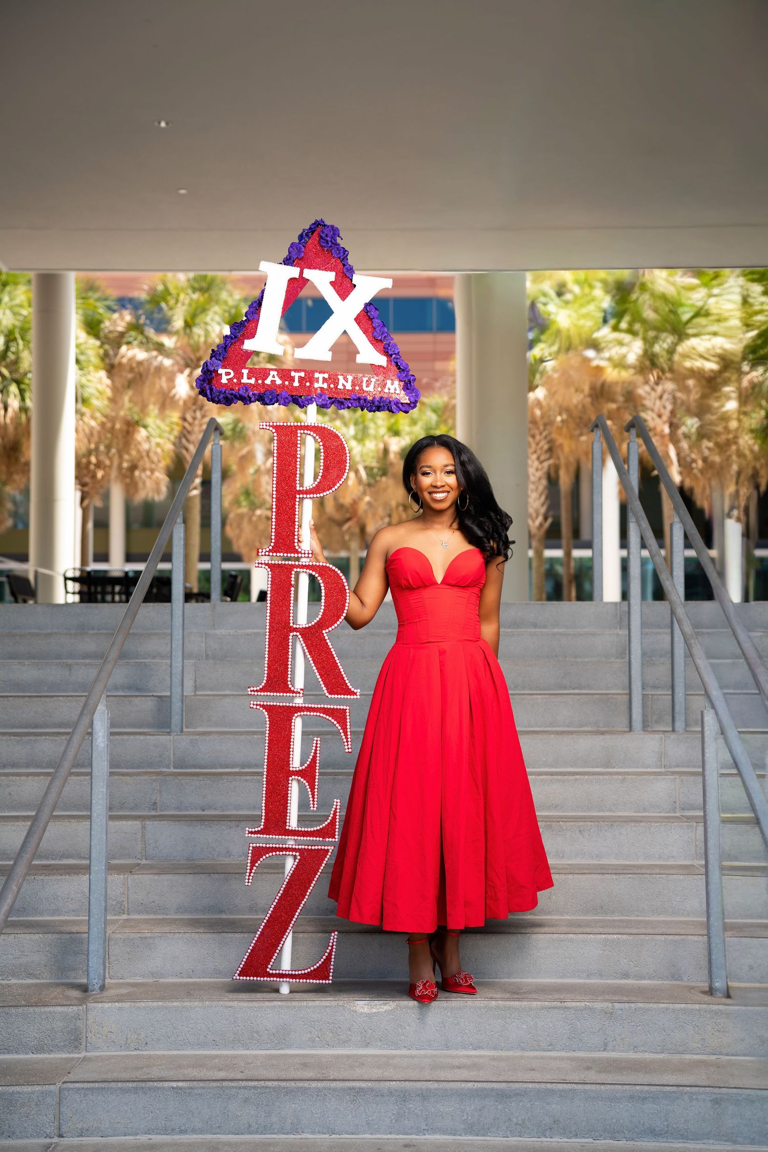 Woman in red strapless midi dress posing beside a decorative PREZ sign on outdoor steps with palm trees.