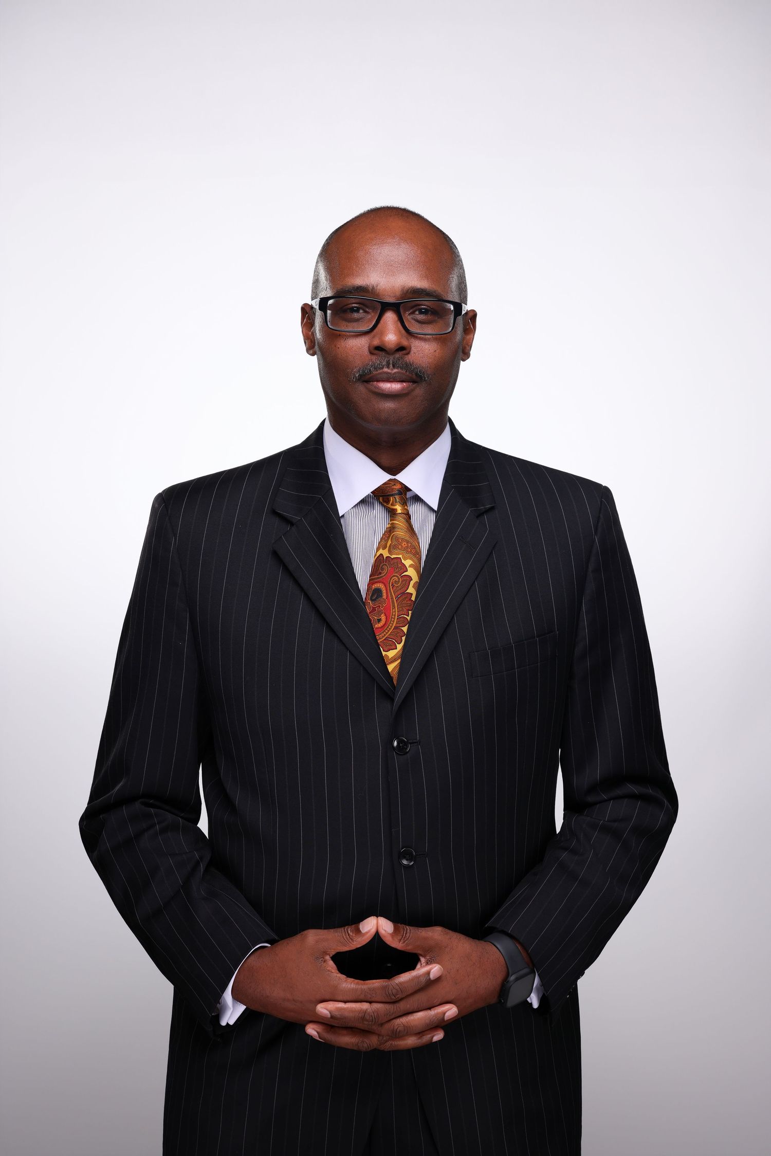 Professional Black businessman in pinstripe suit and orange tie, wearing glasses, hands clasped, studio portrait.