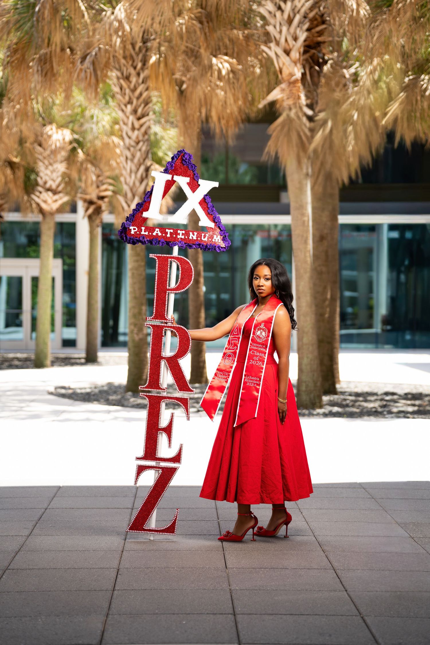 Woman in red dress and sash poses beside illuminated IX Platinum Prez sign outdoors near palm trees.
