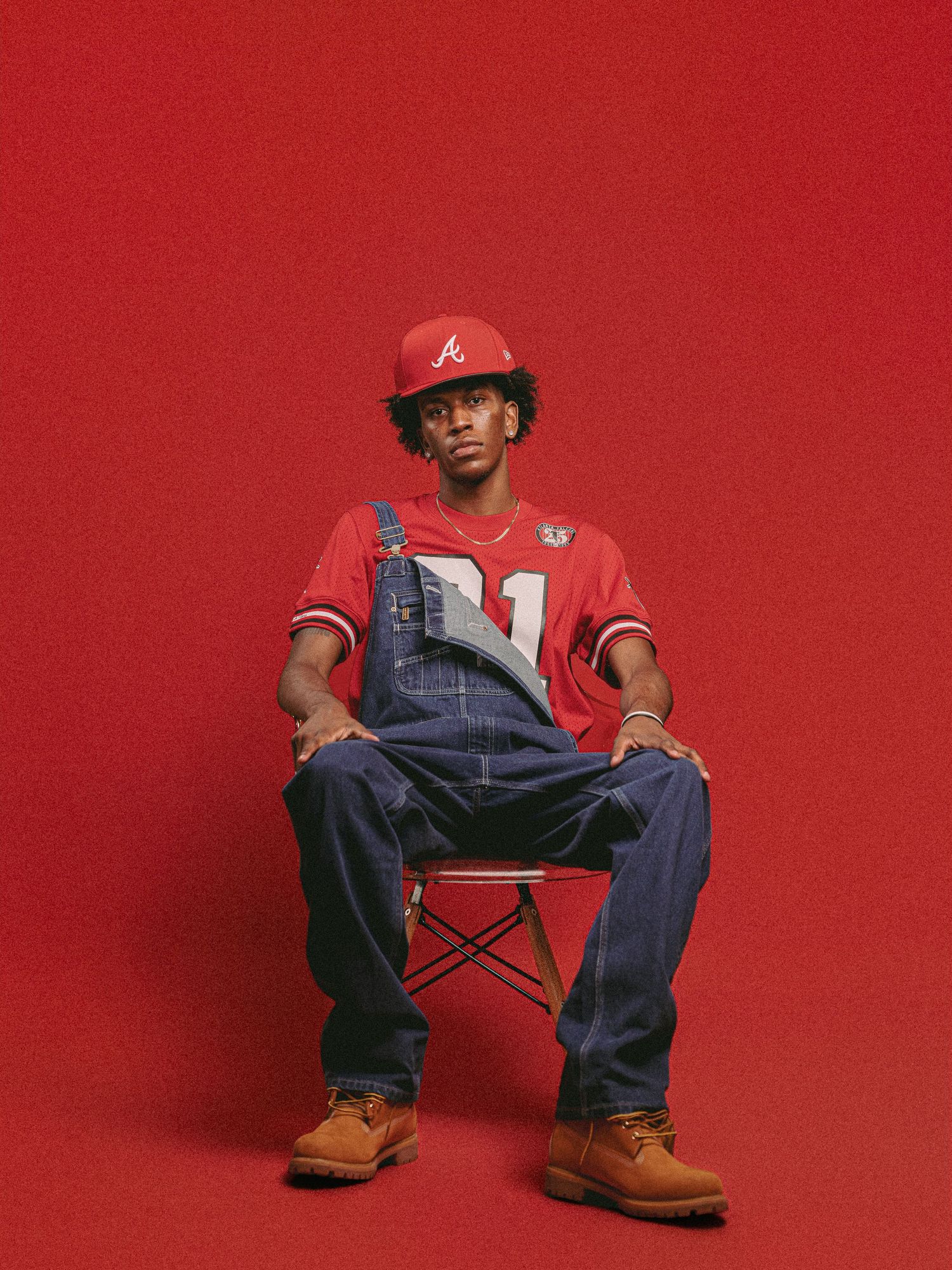 Figure in red baseball cap and denim overalls sits on red folding chair against vibrant red background.
