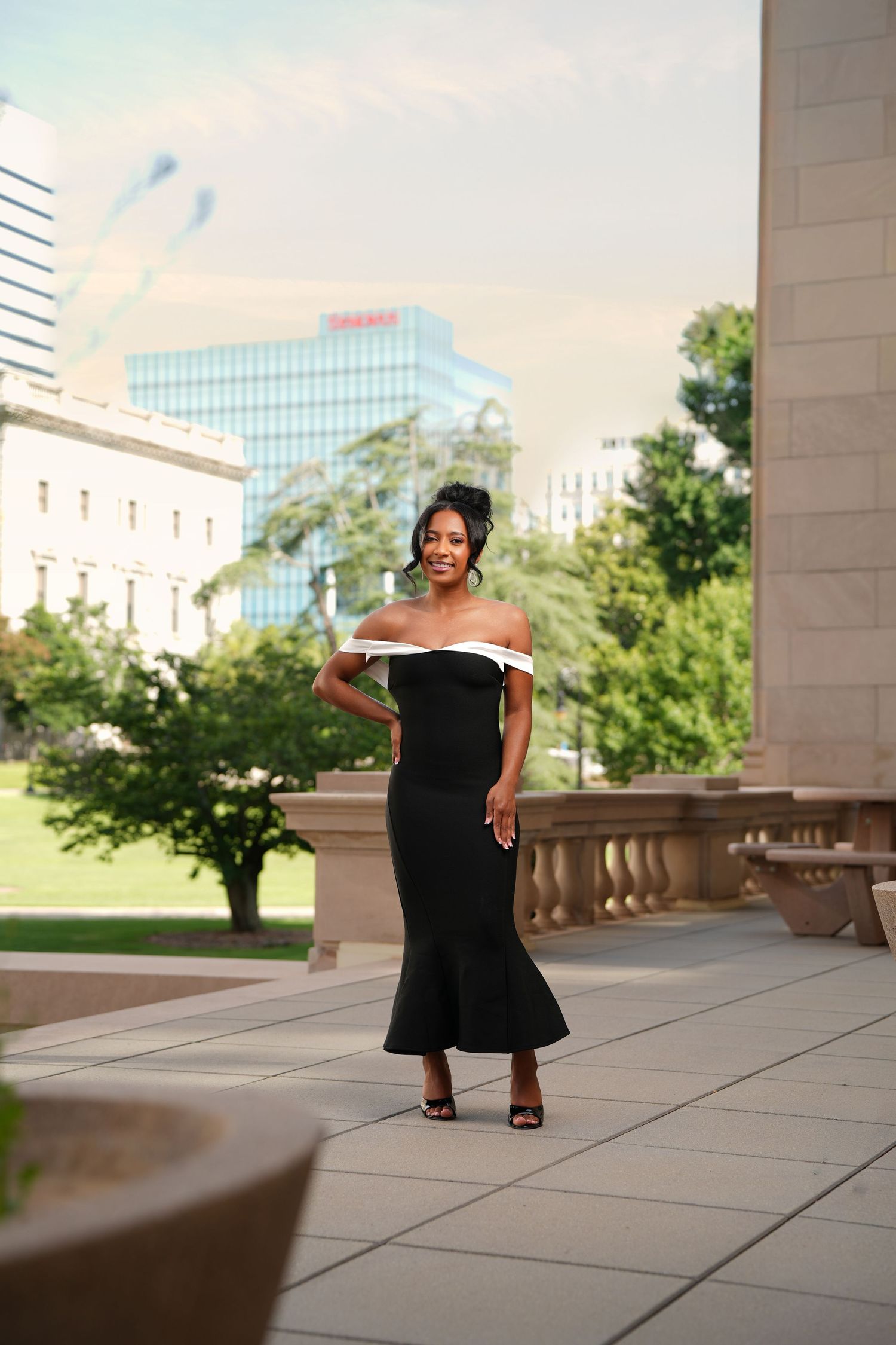 Elegant woman in black off-shoulder mermaid gown posing outdoors near classical architecture and city skyline.