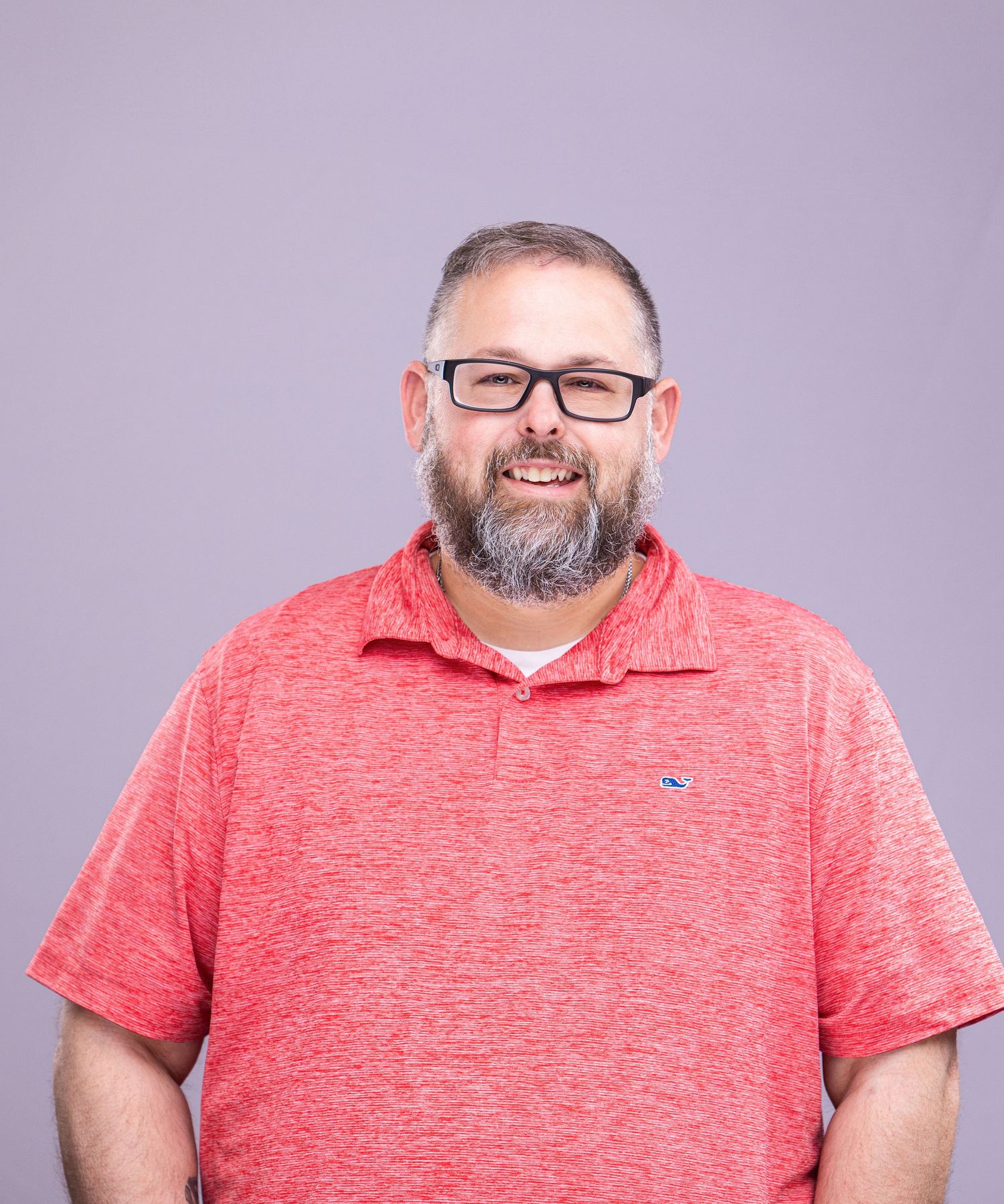 Professional headshot of person wearing glasses and coral polo shirt against purple background.