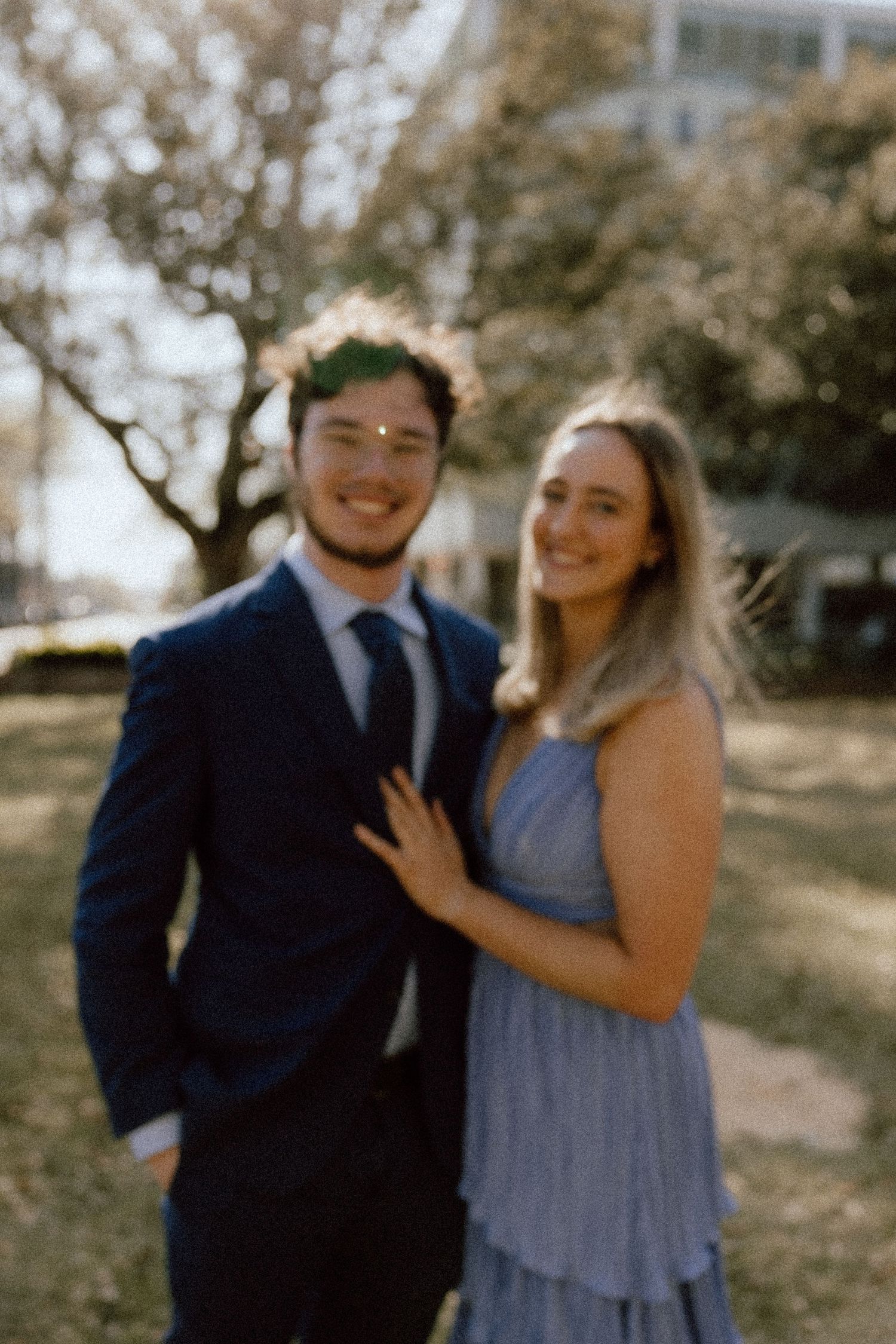 A couple poses outdoors in formal attire, with him in a navy suit and her in a blue dress.