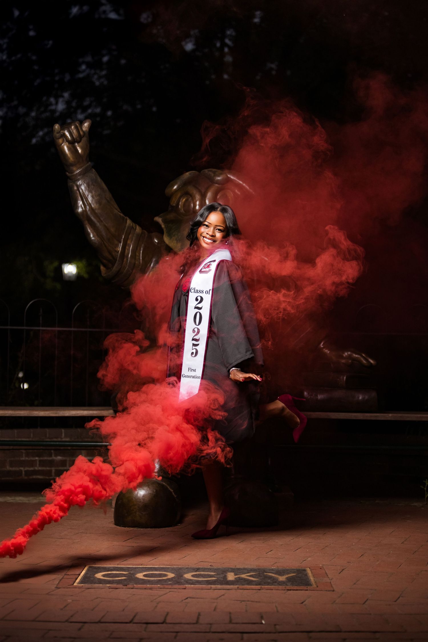 Graduate poses with mascot statue amid dramatic red smoke effects.