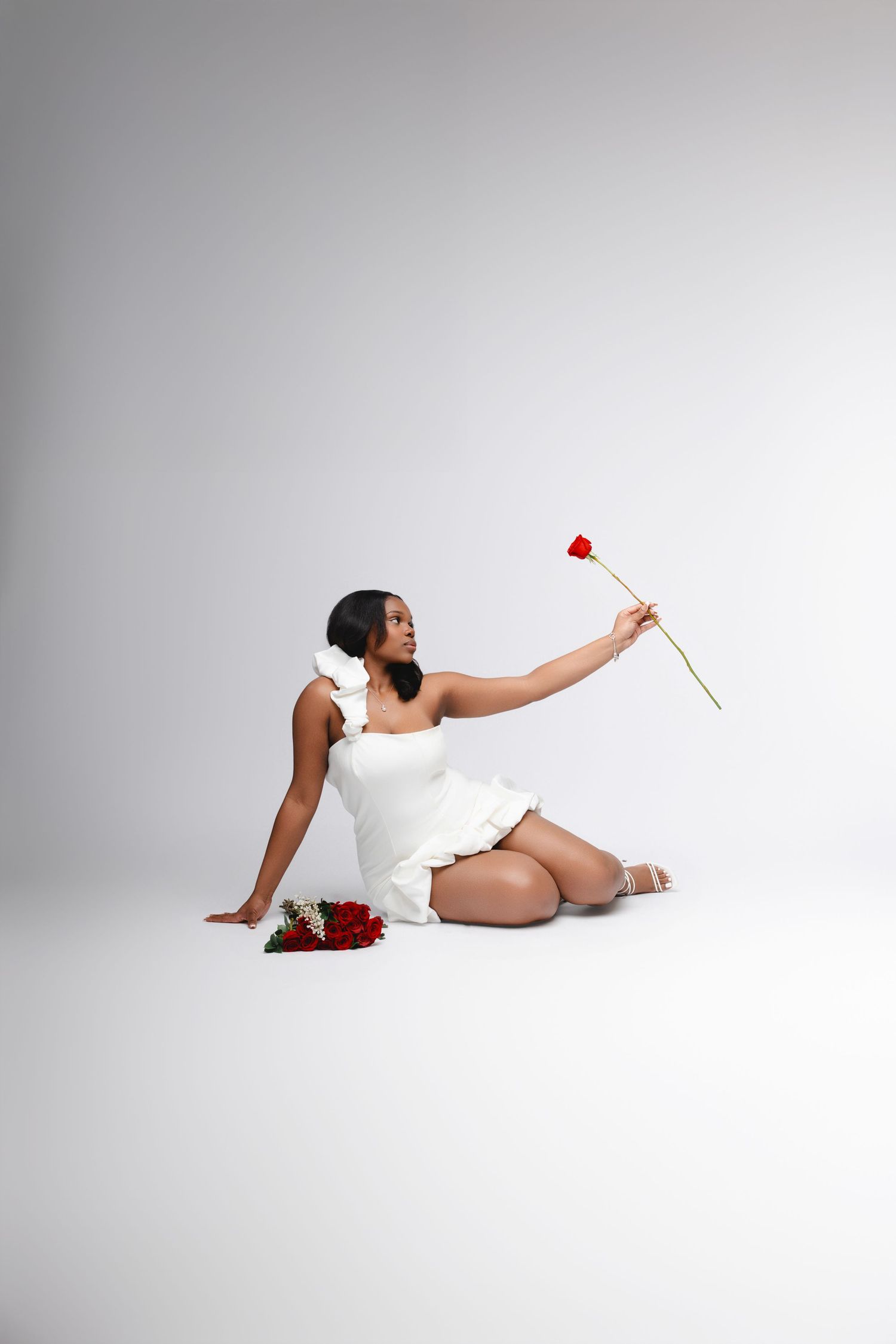 Woman in white dress poses elegantly with red roses on studio floor.