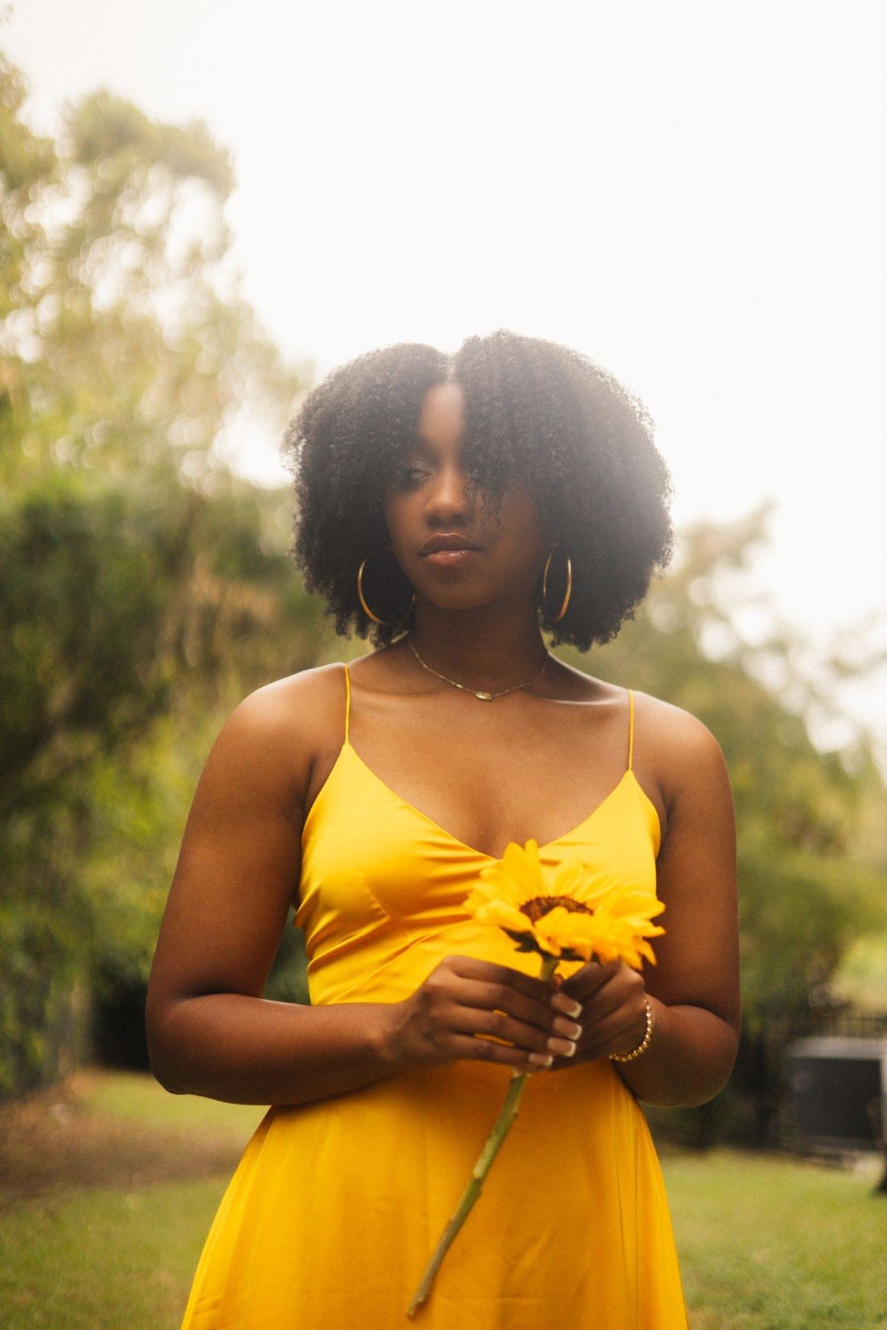 Person in bright yellow sundress holds a yellow flower in an outdoor garden setting during golden hour.
