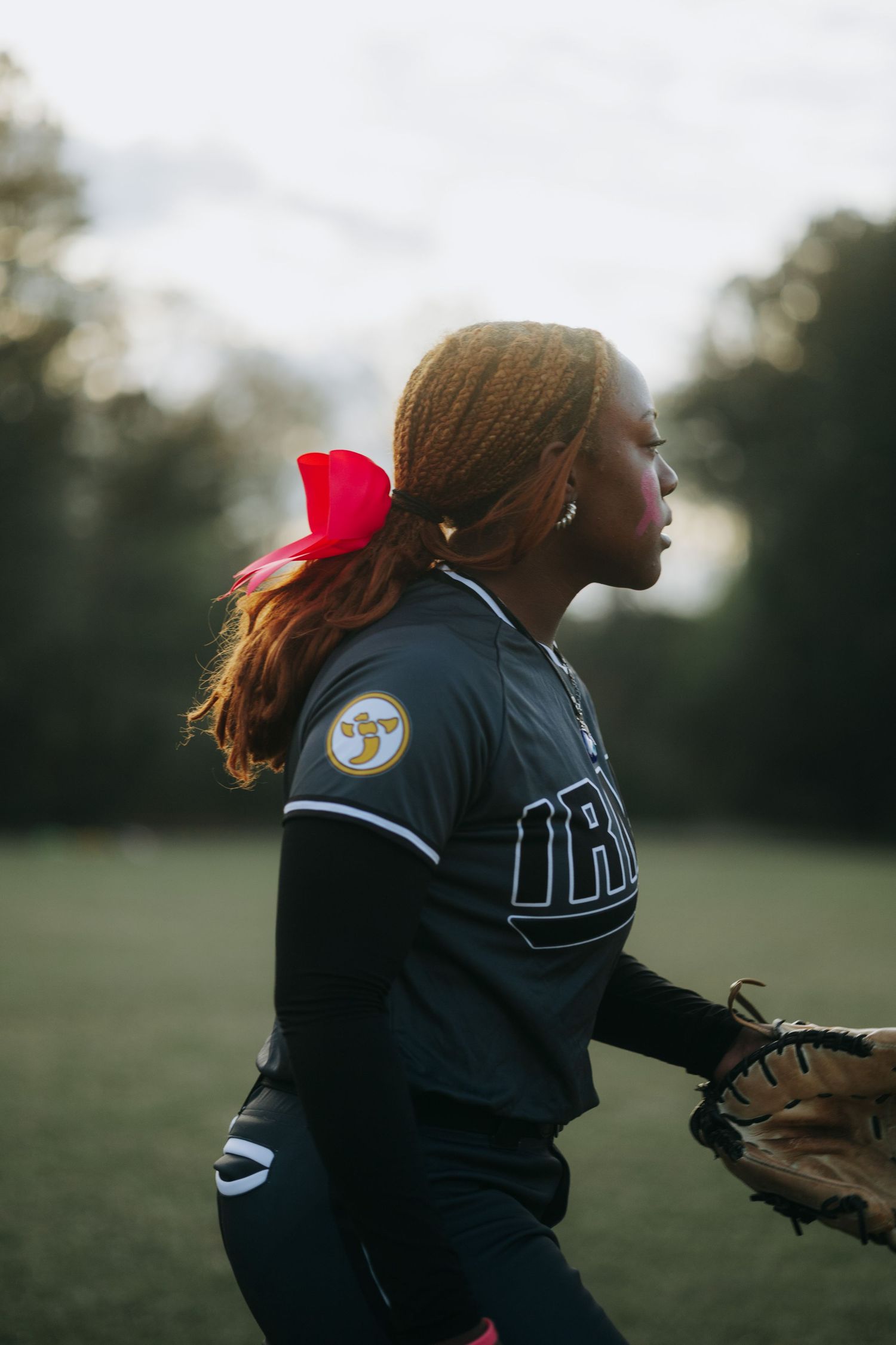 Irmo HS Softball player wearing a dark uniform with a red bow in their hair stands ready with a glove on the field.