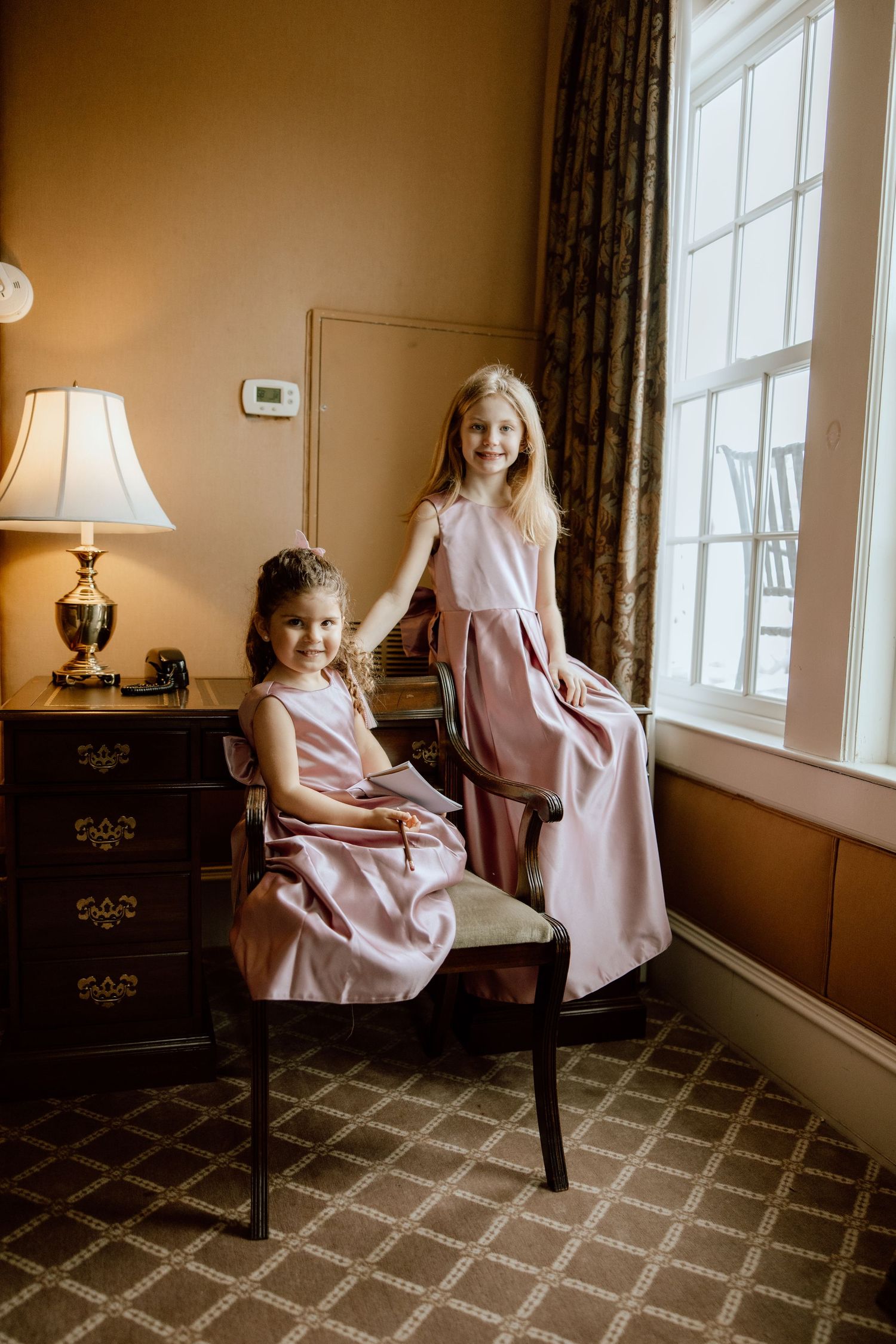 Two children in matching pink dresses pose playfully in a sunlit room with patterned carpet and traditional decor.