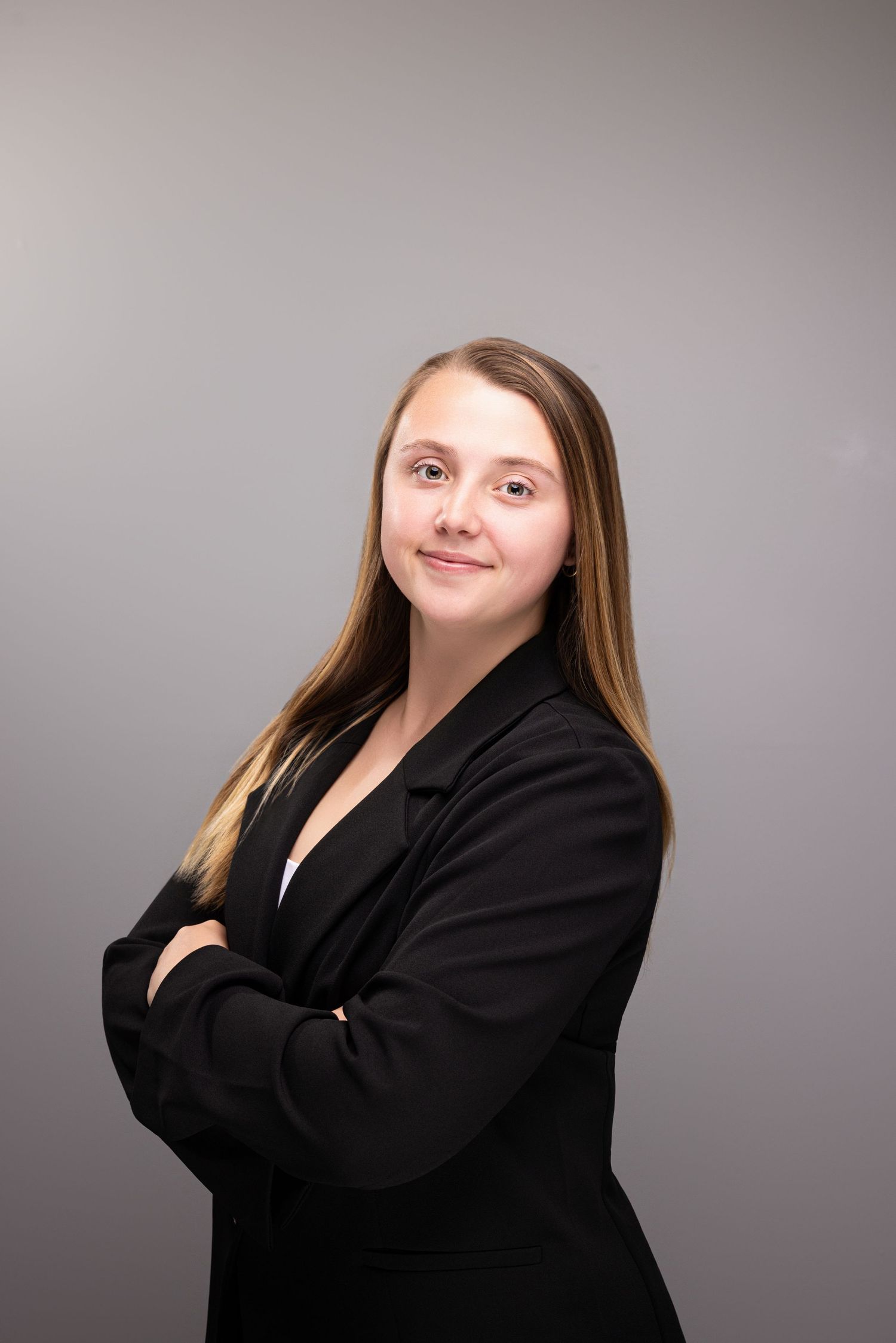 Young professional woman in black blazer with arms crossed, smiling confidently against gray background.