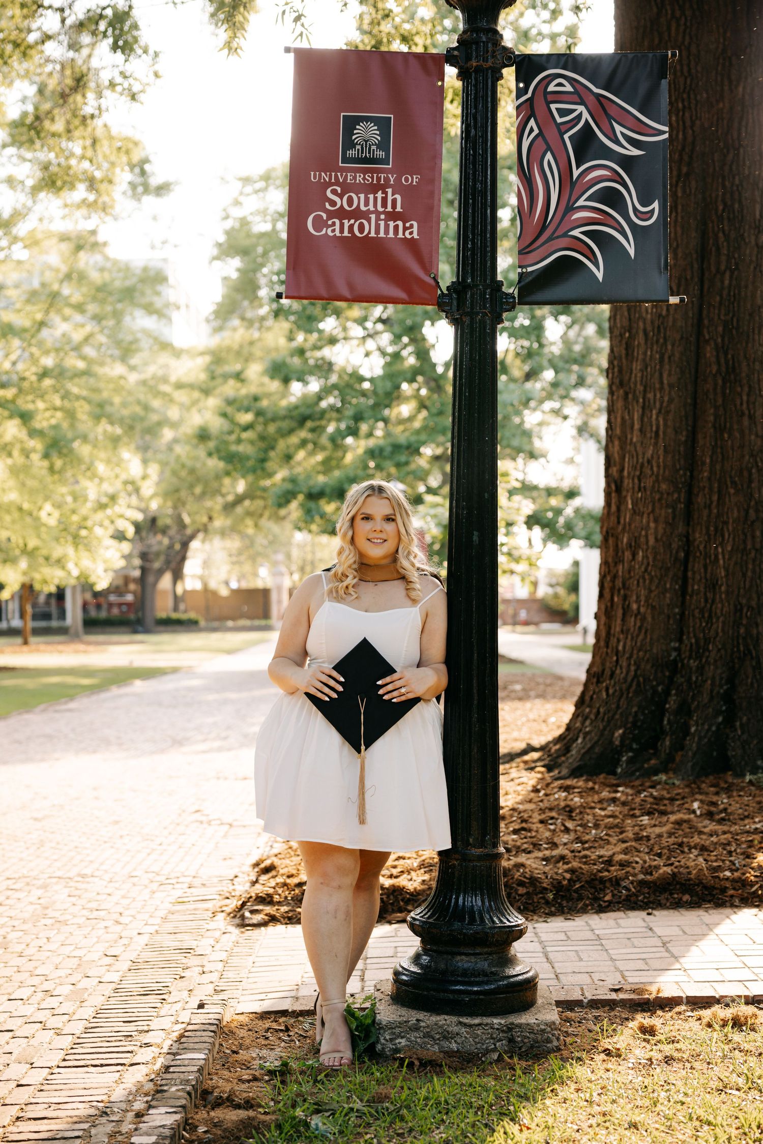 Graduate in white dress poses by University of South Carolina banner holding cap.