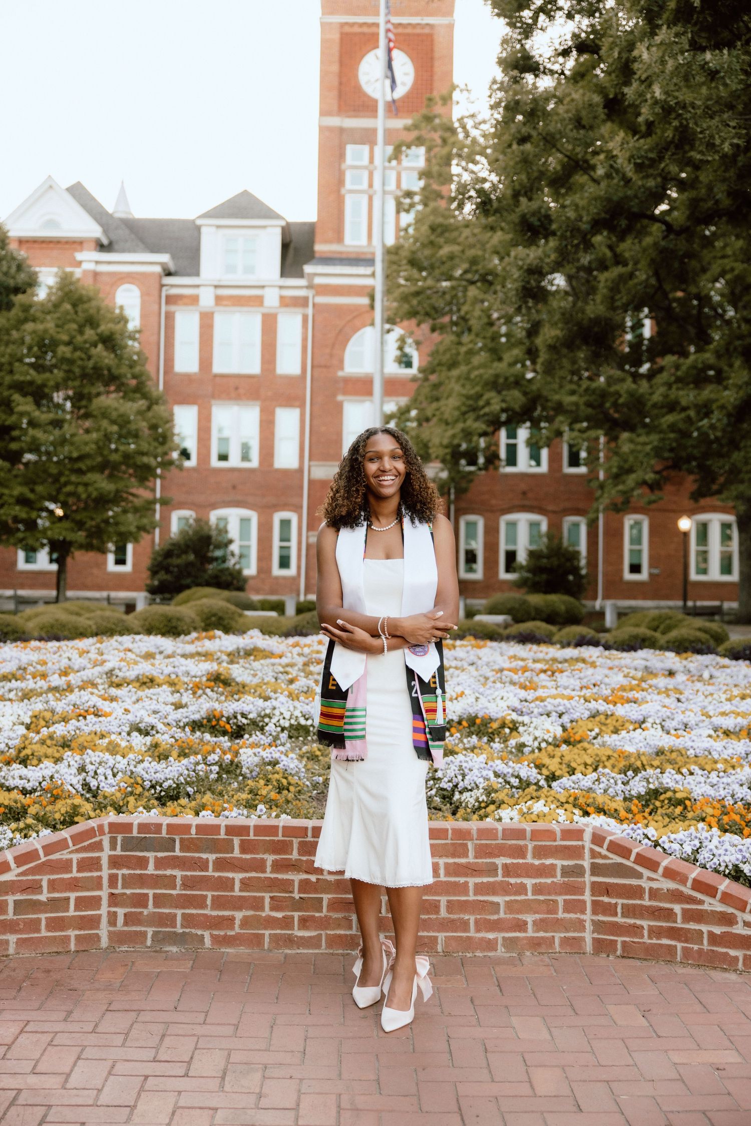 Young woman in white dress and kente stole poses by flower garden at university campus graduation photo.