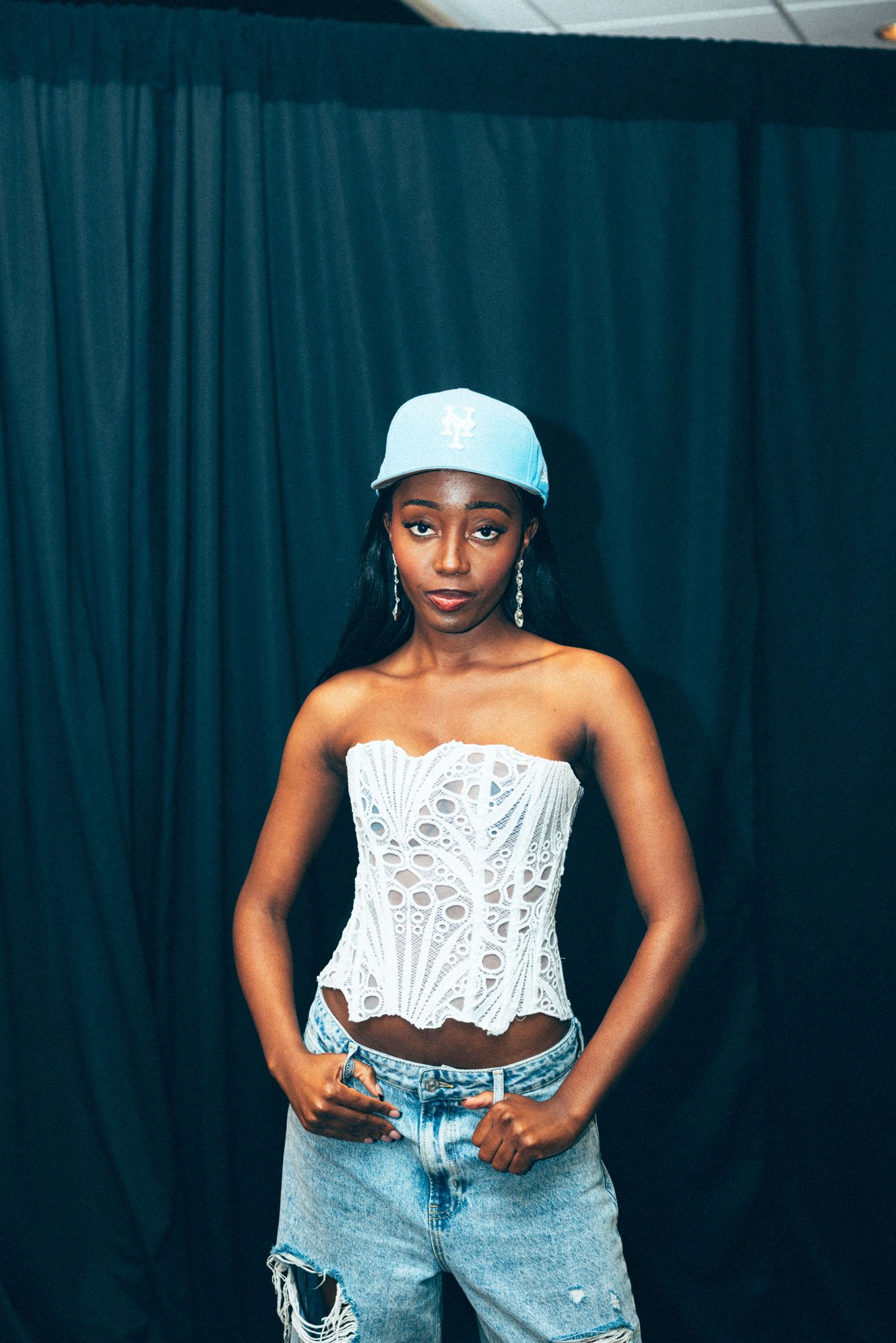 Young Black woman in white corset top, blue cap, and ripped jeans posing confidently against dark teal curtain backdrop.