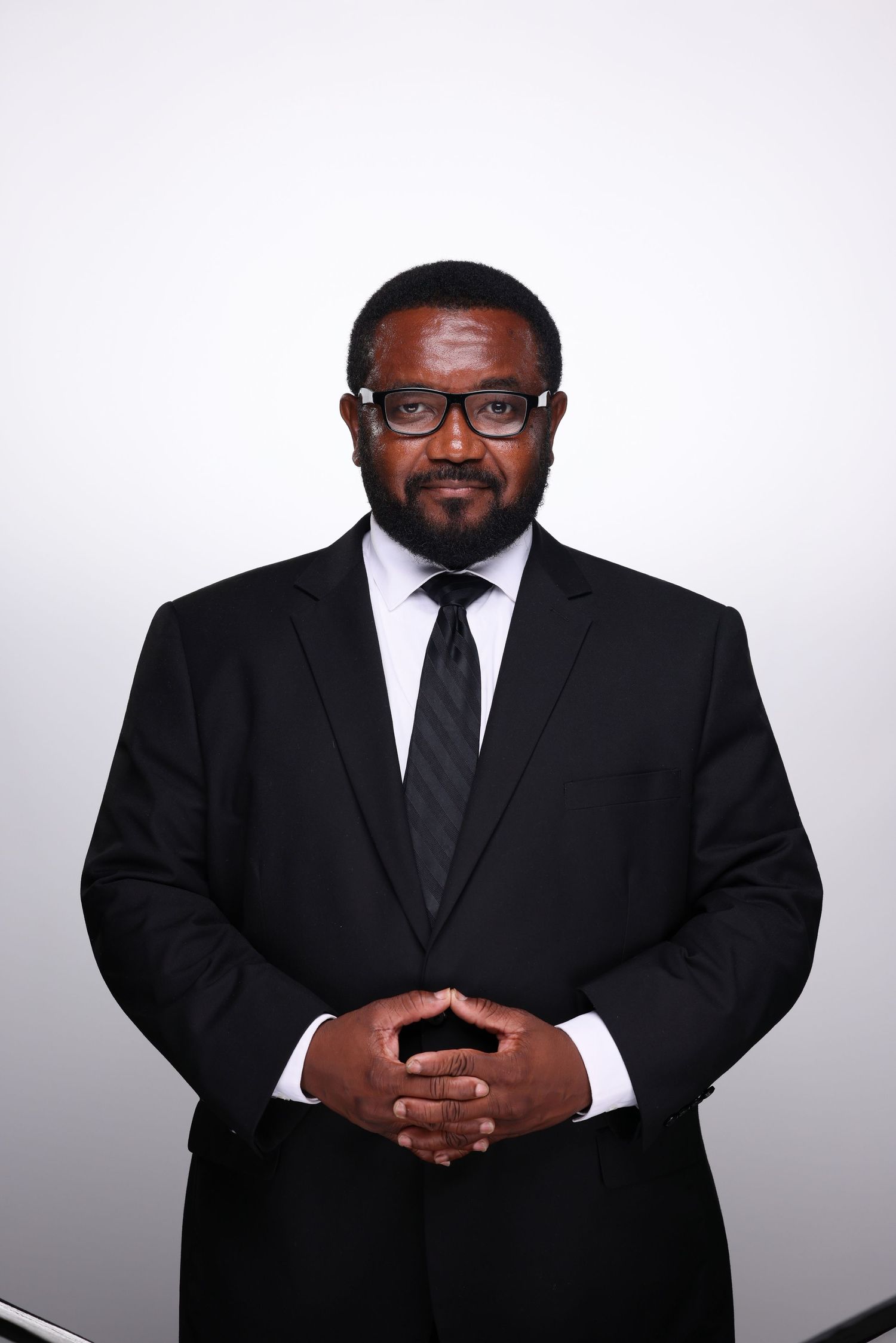 Professional Black man in black suit, white shirt, and tie with glasses, posing confidently with hands clasped.
