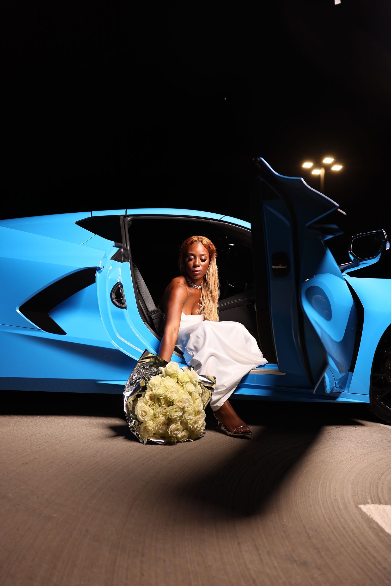 A blue sports car with a person holding a white floral bouquet, photographed from a low angle against a beige floor.