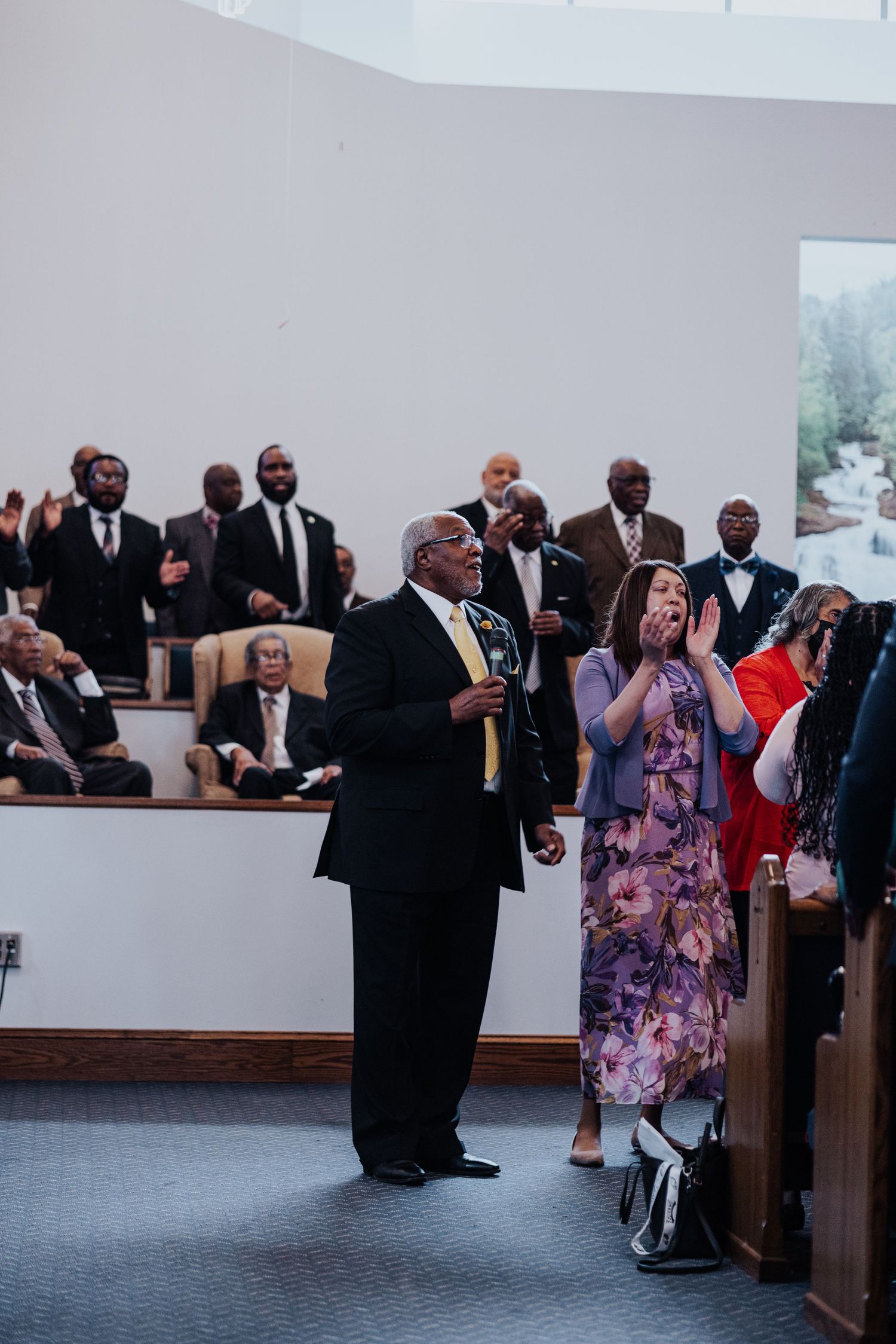 Wedding guests stand in church pews during a ceremony, some wearing formal attire and others in colorful dresses.