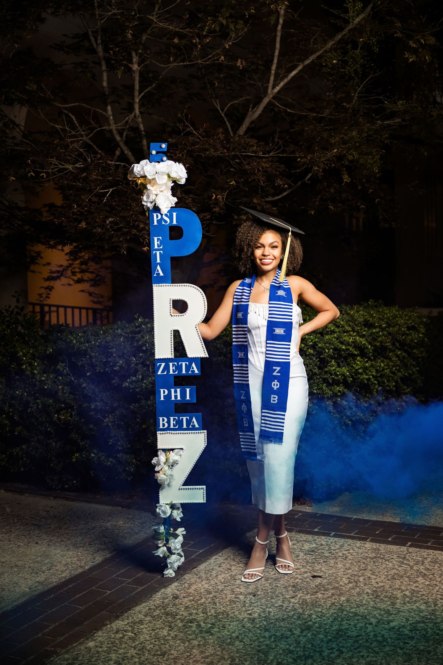 Graduate in cap and gown poses beside Zeta Phi Beta sorority sign with blue smoke at night.