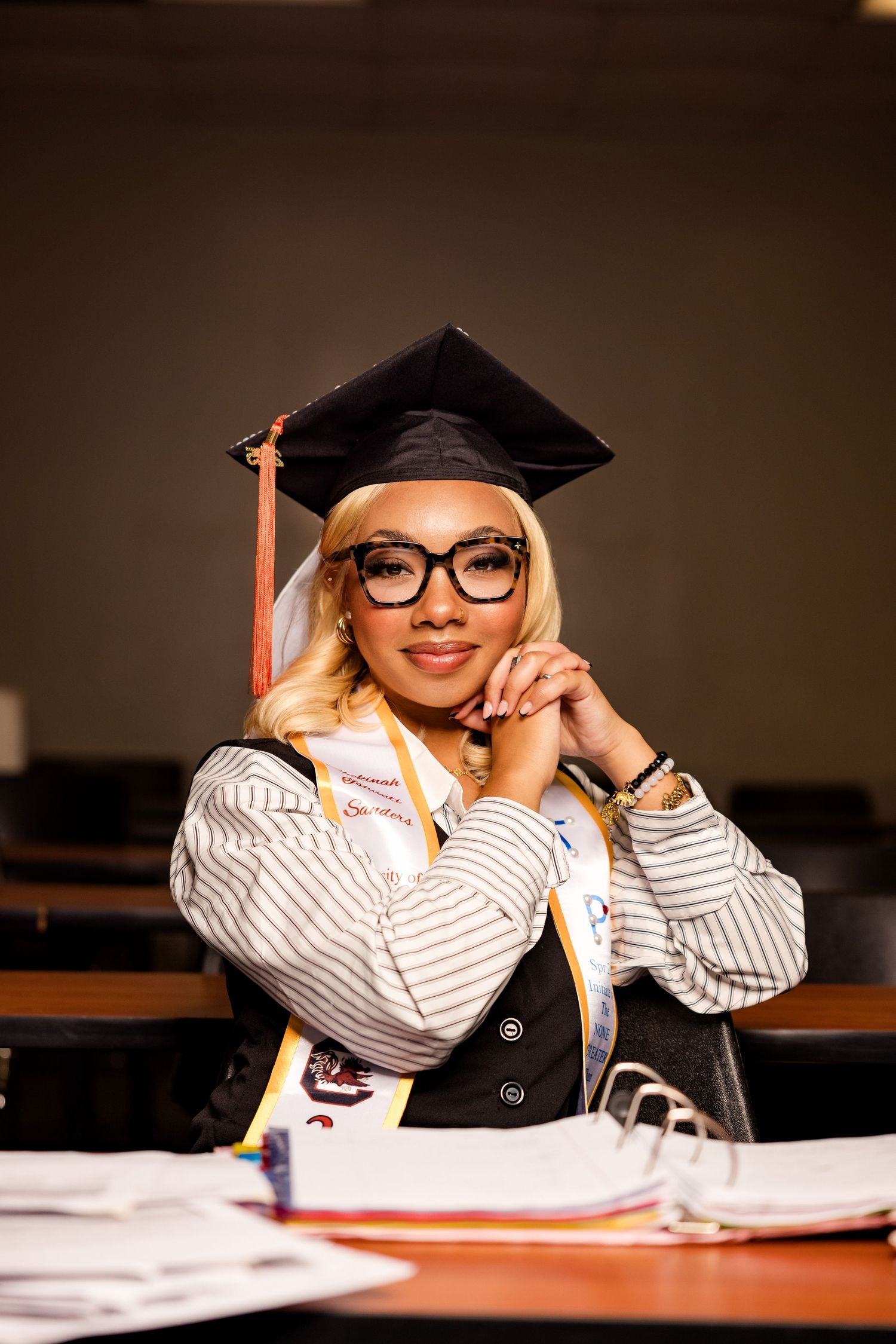 Graduate in cap and gown smiles while sitting at desk with study materials.