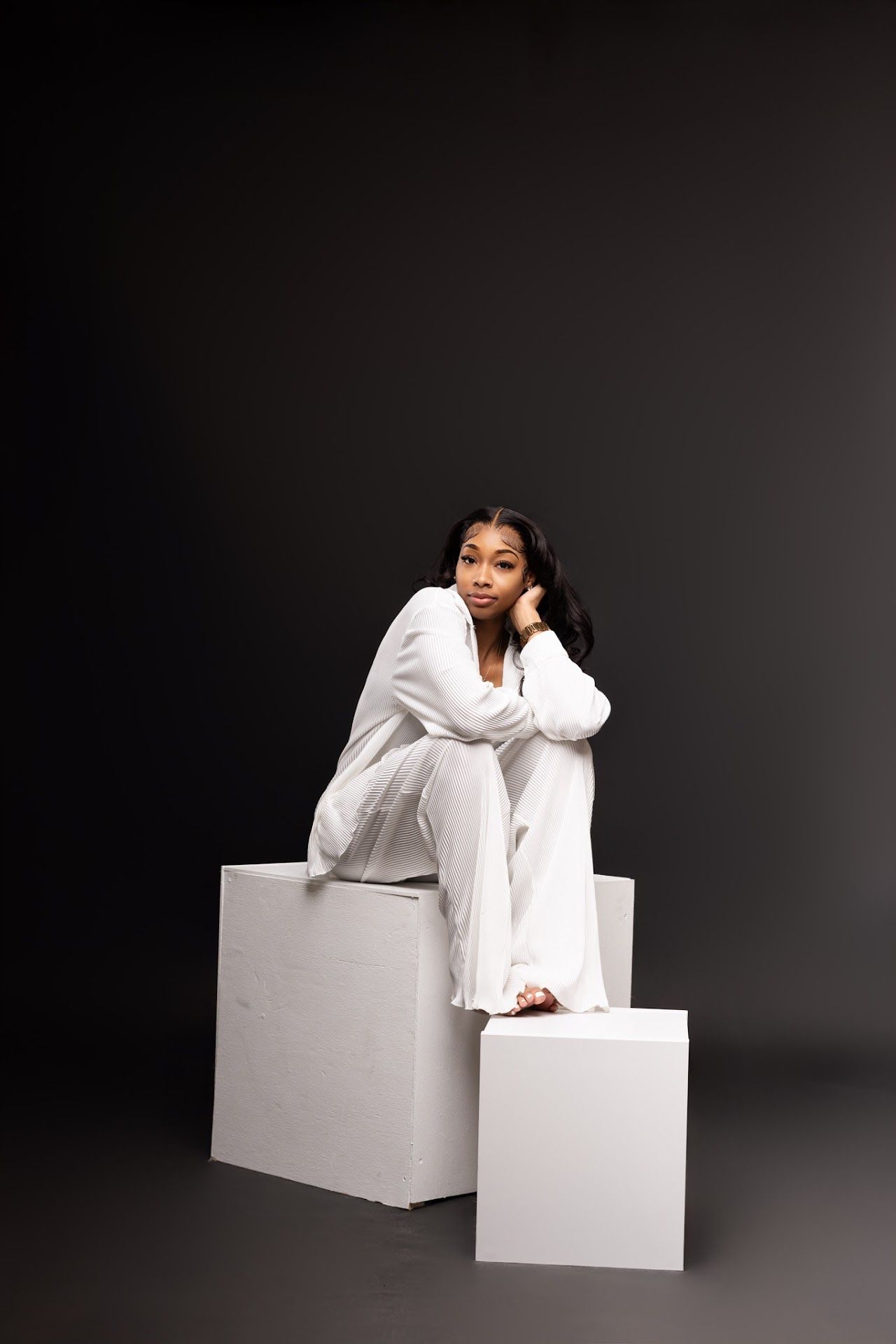 Person in an all-white outfit sits elegantly on white geometric blocks against a dark background in a studio setting.