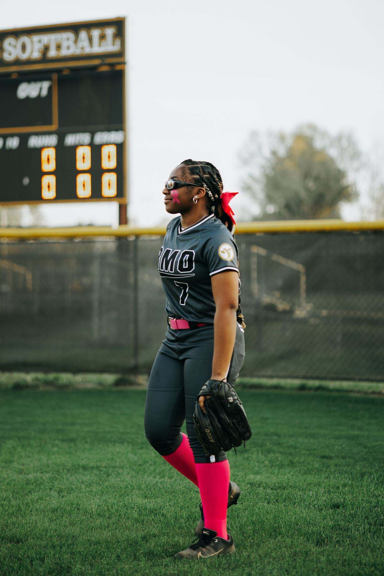 A Irmo HS softball player in dark uniform with pink socks stands ready on the field with scoreboard visible.