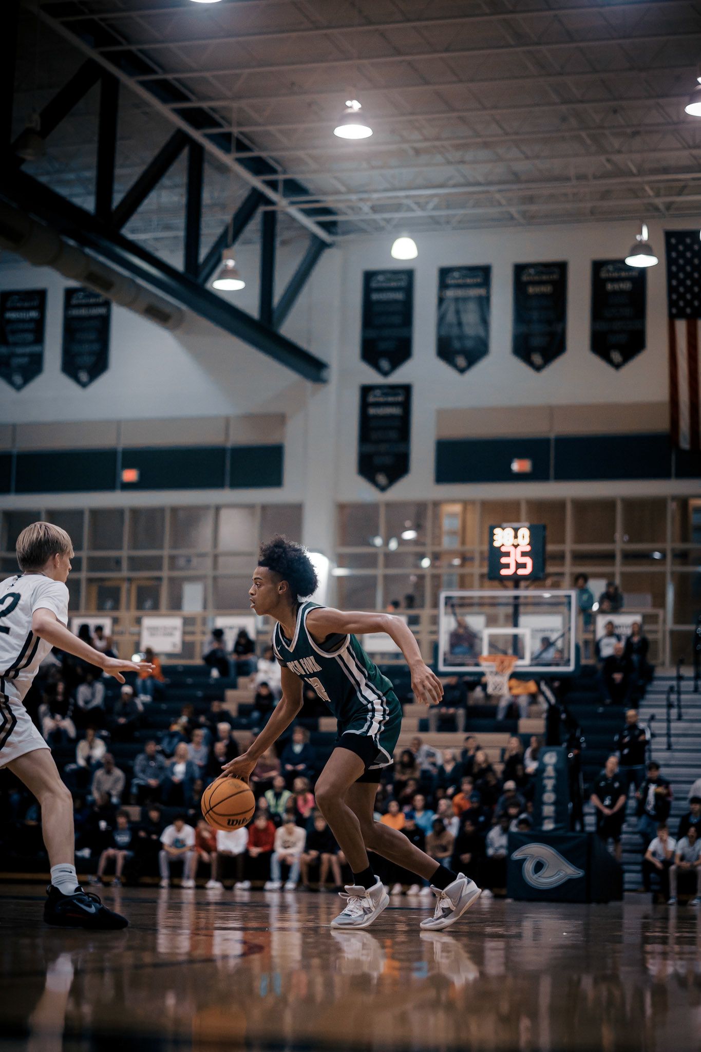 Basketball player dribbles past defender during indoor game with championship banners visible.