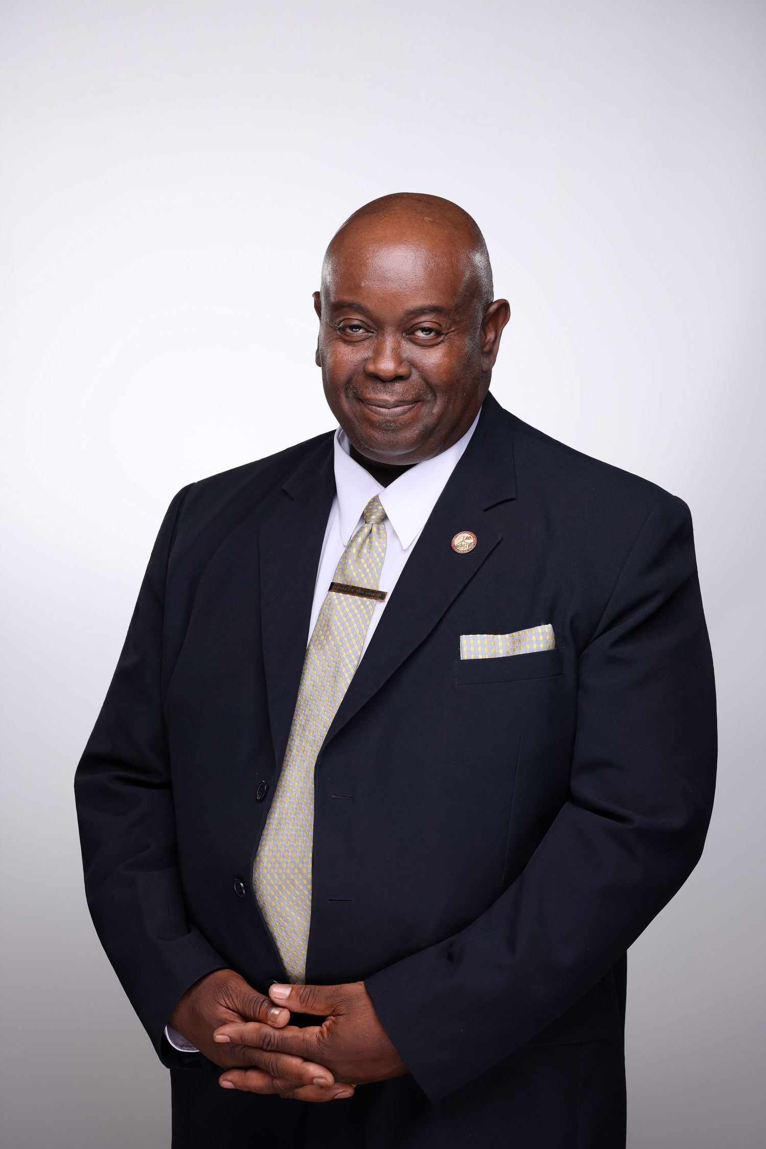Professional man in dark suit and gold tie smiling, wearing lapel pin, hands clasped, formal portrait photo.