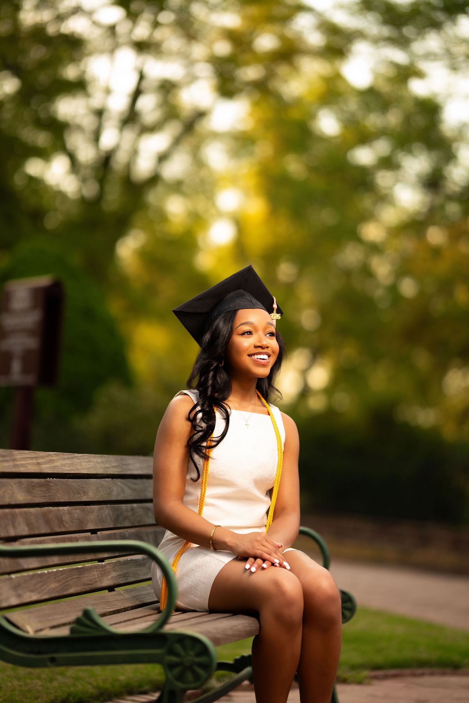 Smiling young woman in graduation cap and yellow sash sits on a park bench wearing a white dress.