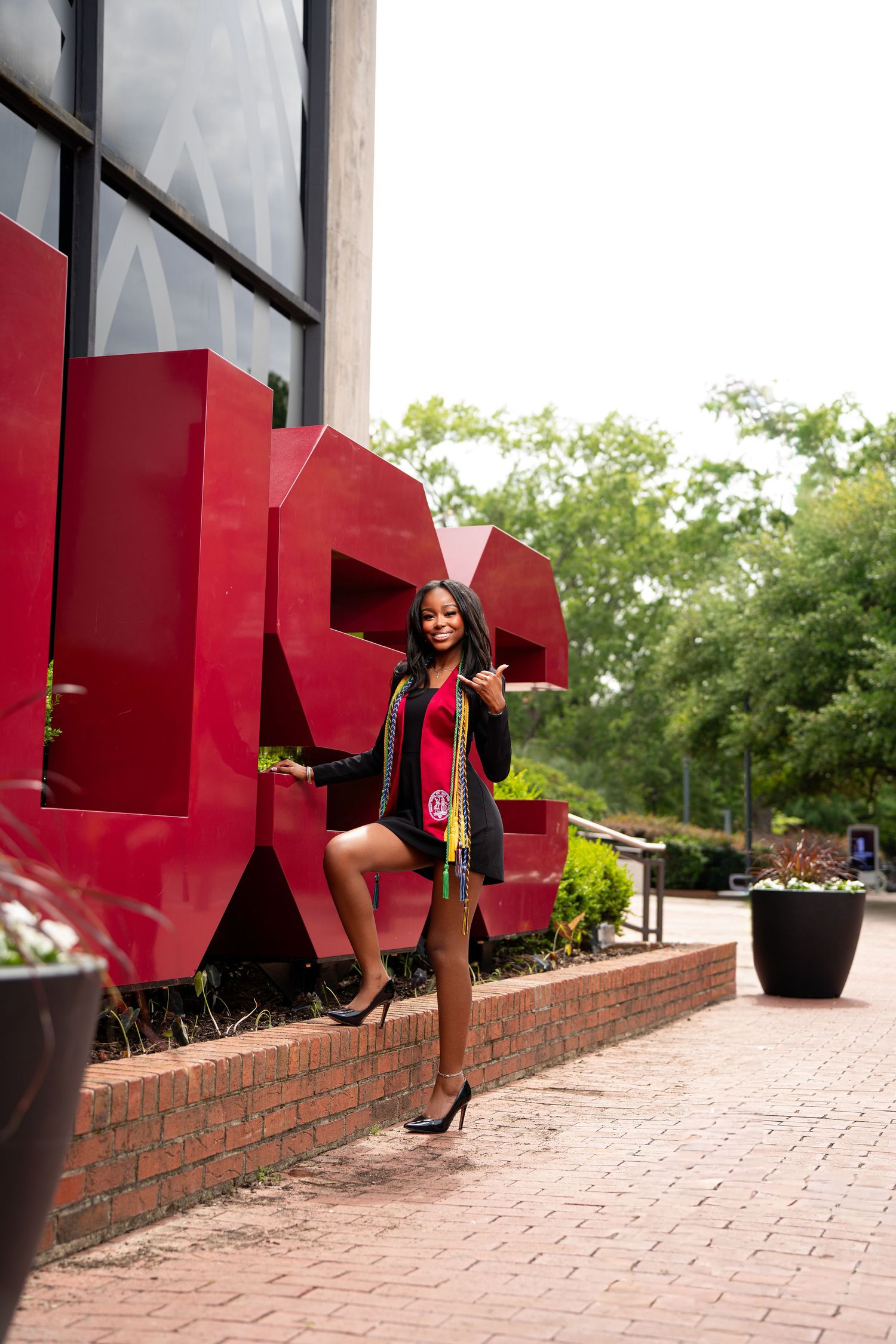 A graduate poses in cap and gown with red scarf before large red USG letters on a brick plaza.