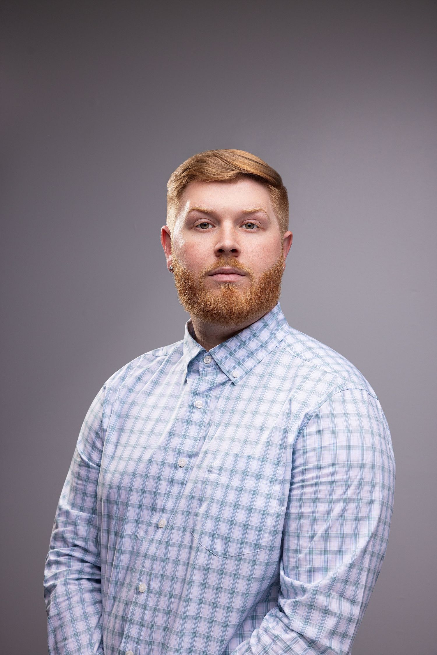 Red-bearded man in blue plaid shirt poses for professional headshot against gray background.