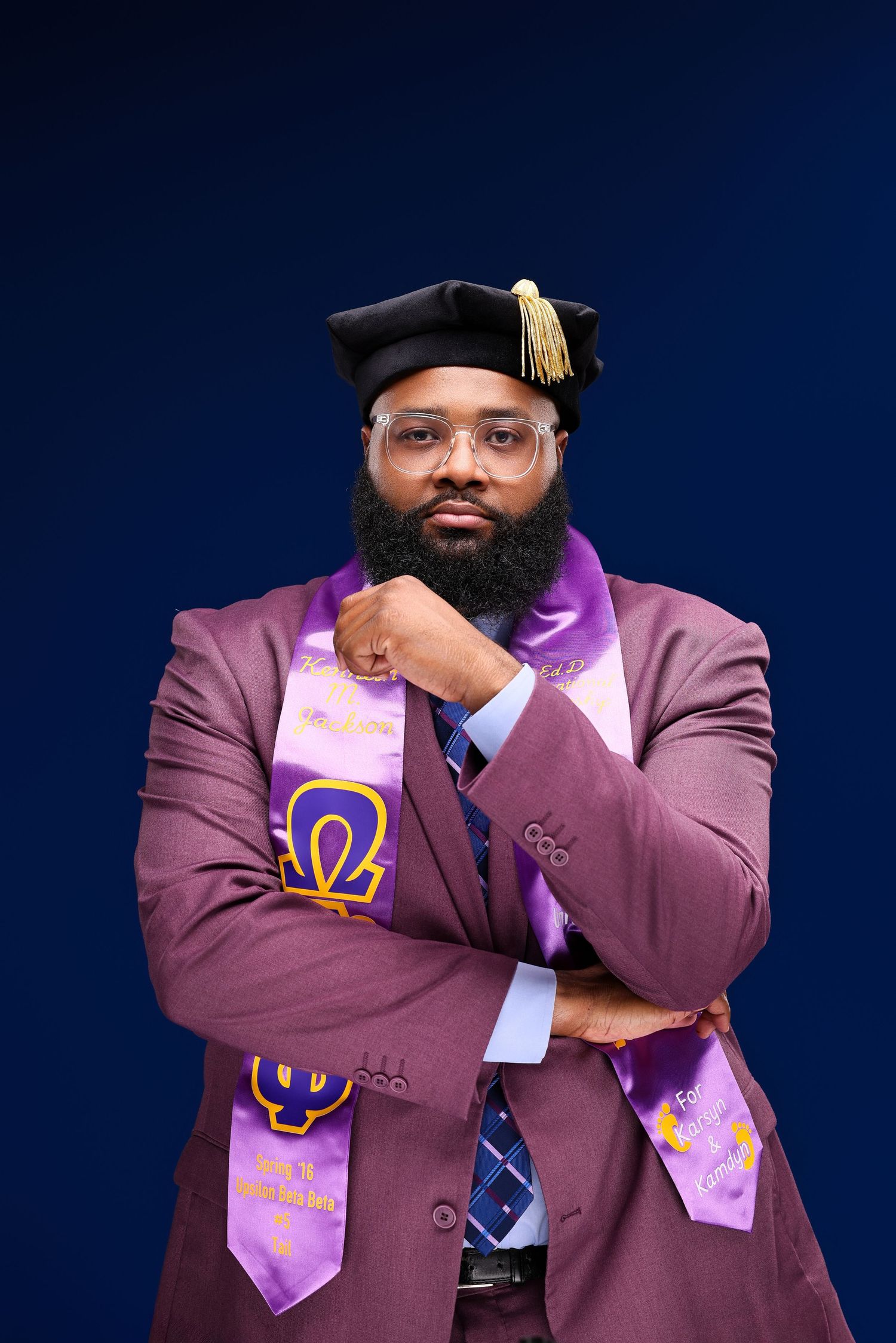 Graduate wearing purple graduation stole and burgundy suit poses with crossed arms against dark blue background.