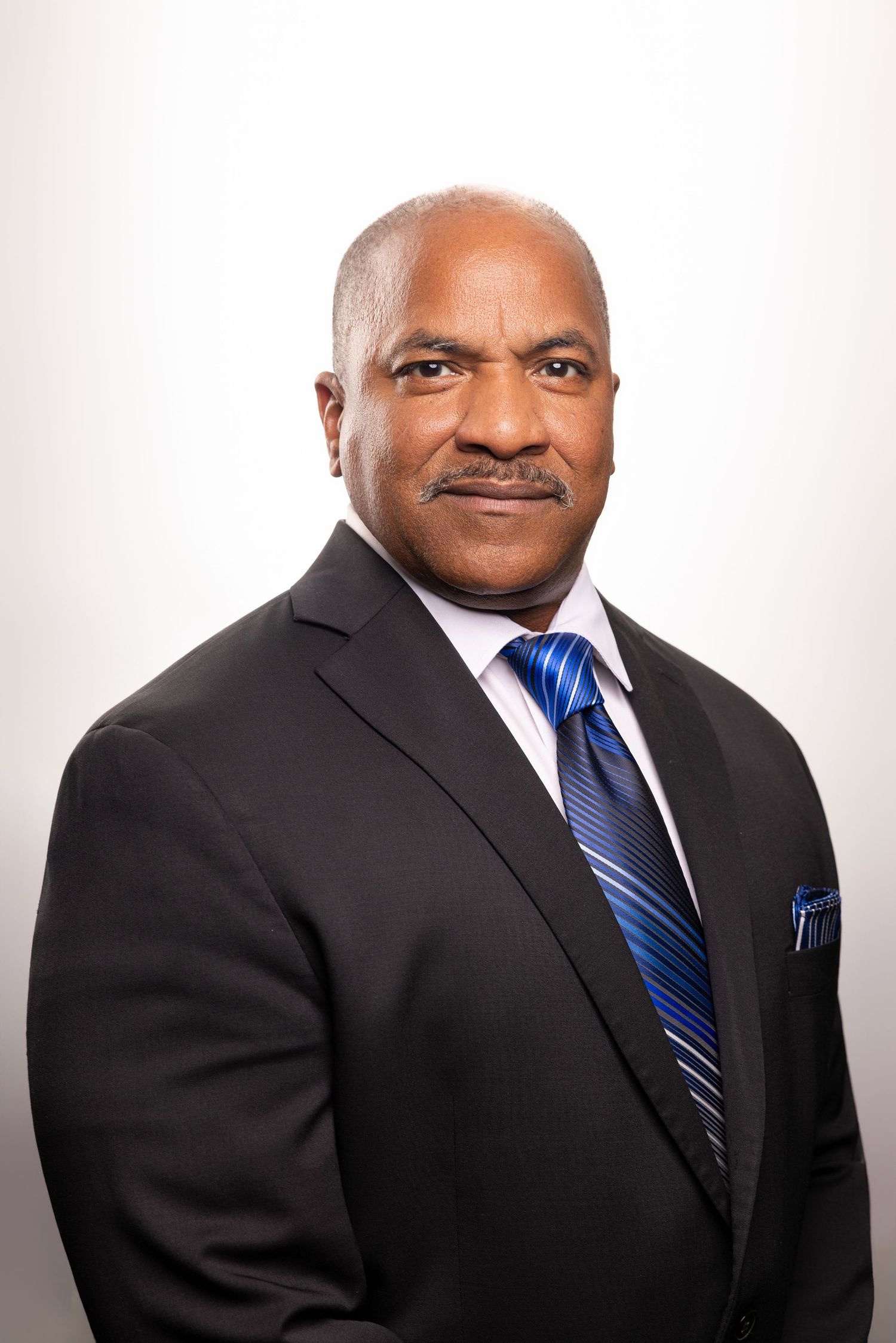 Professional headshot of a man in a black suit with blue striped tie and pocket square on white background.