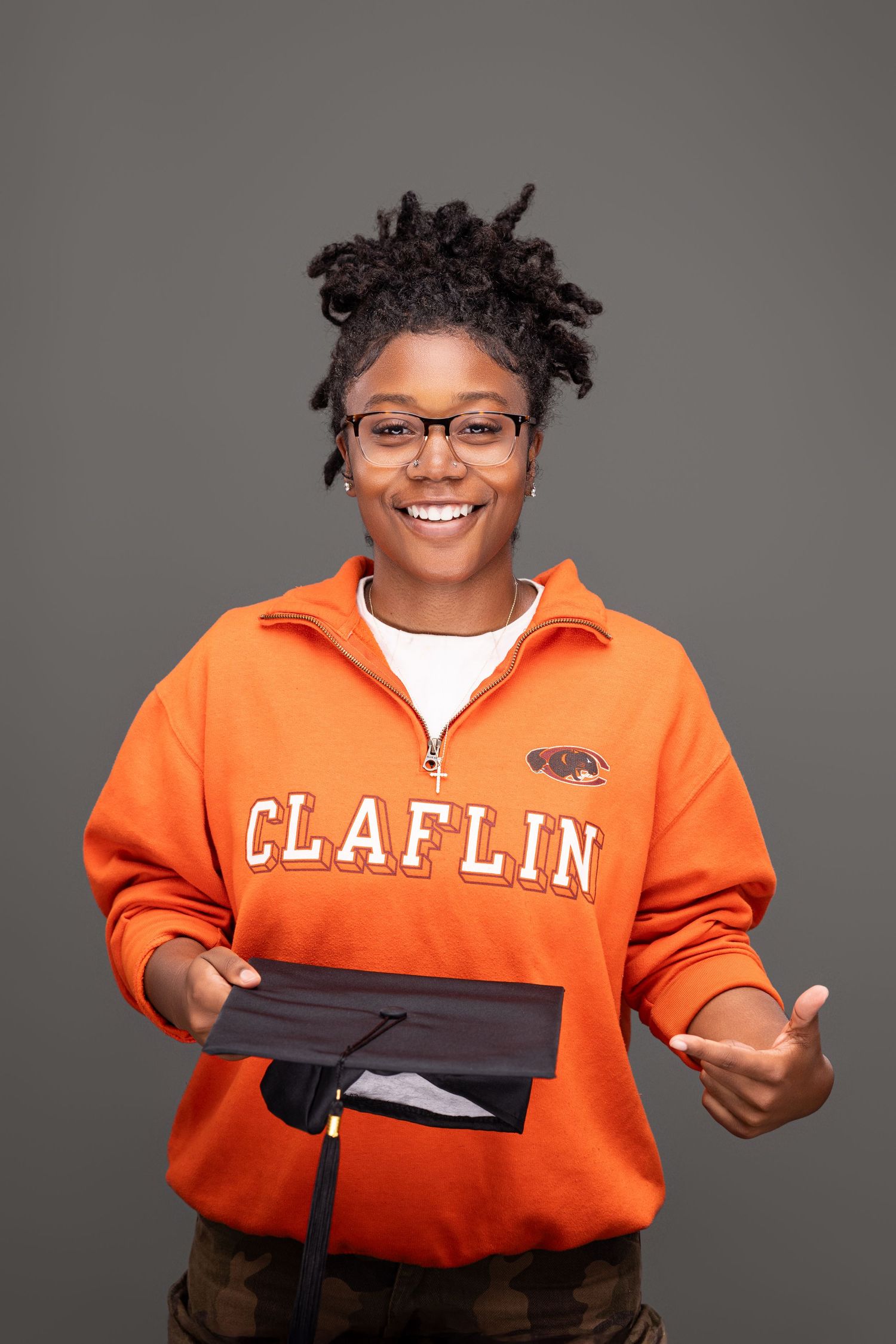 Student wearing orange Claflin University sweatshirt holding graduation cap and smiling.