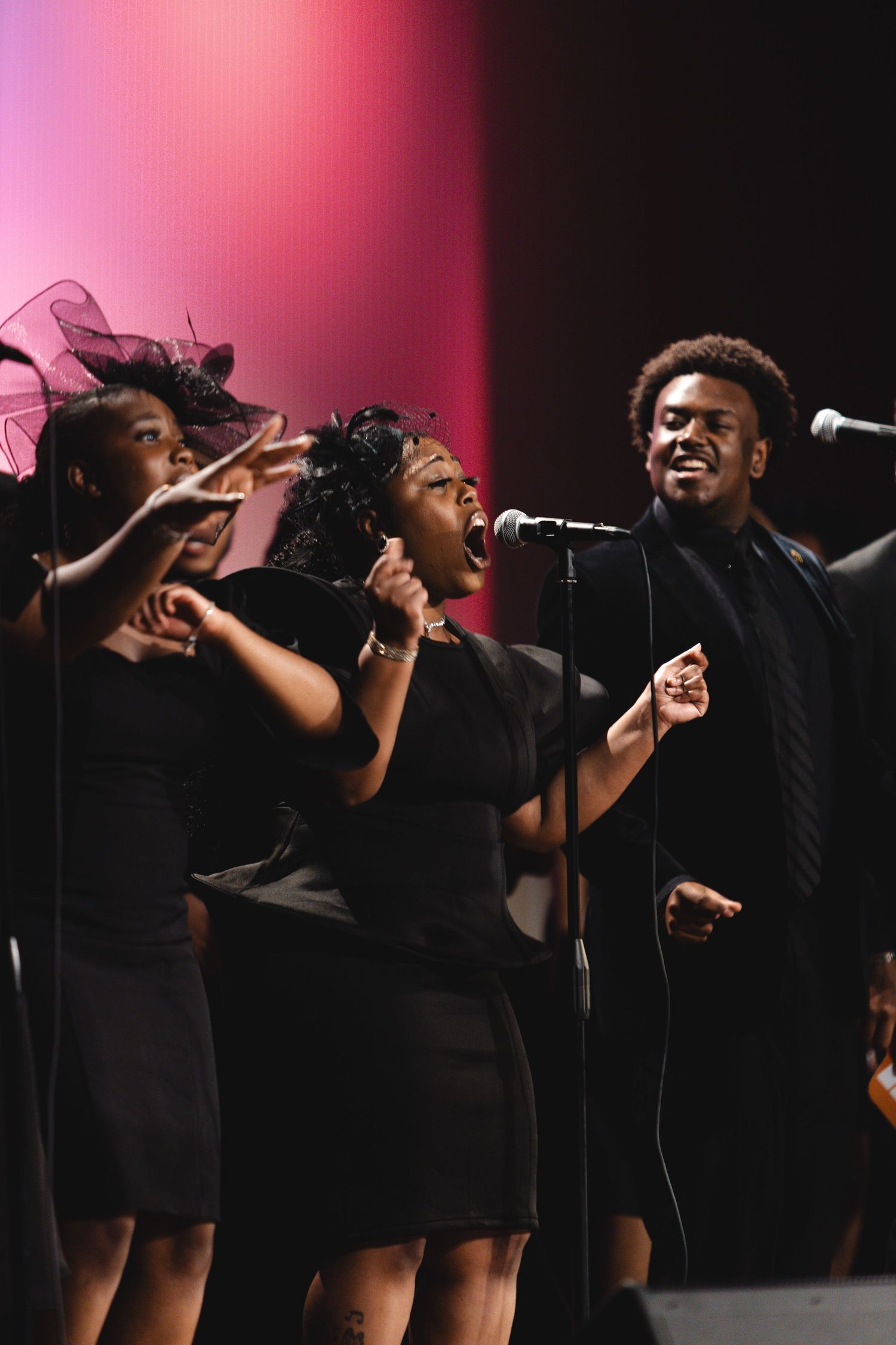 Gospel choir members in black attire performing passionately against pink lighting.
