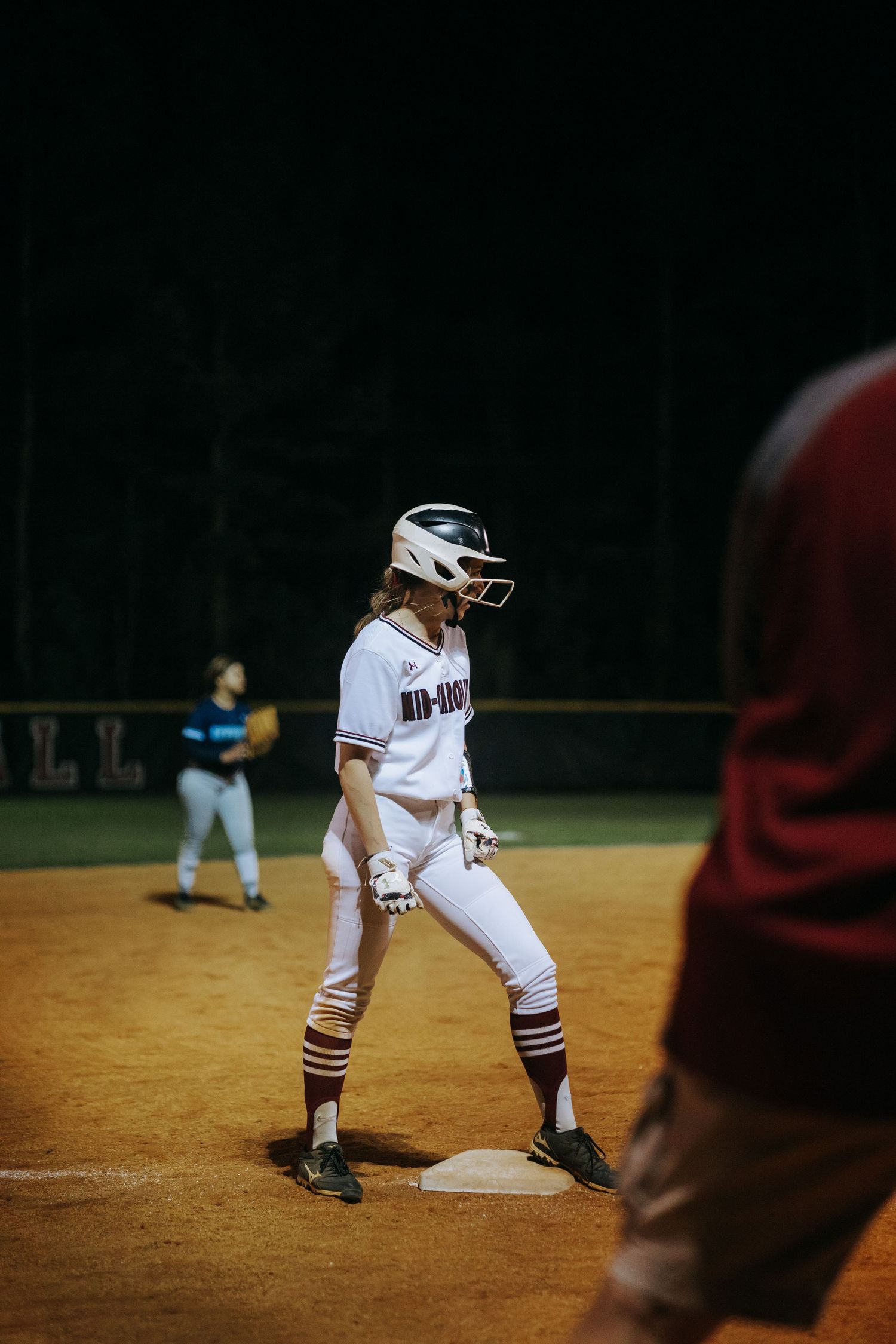 Softball player in white uniform takes lead off base during nighttime game.