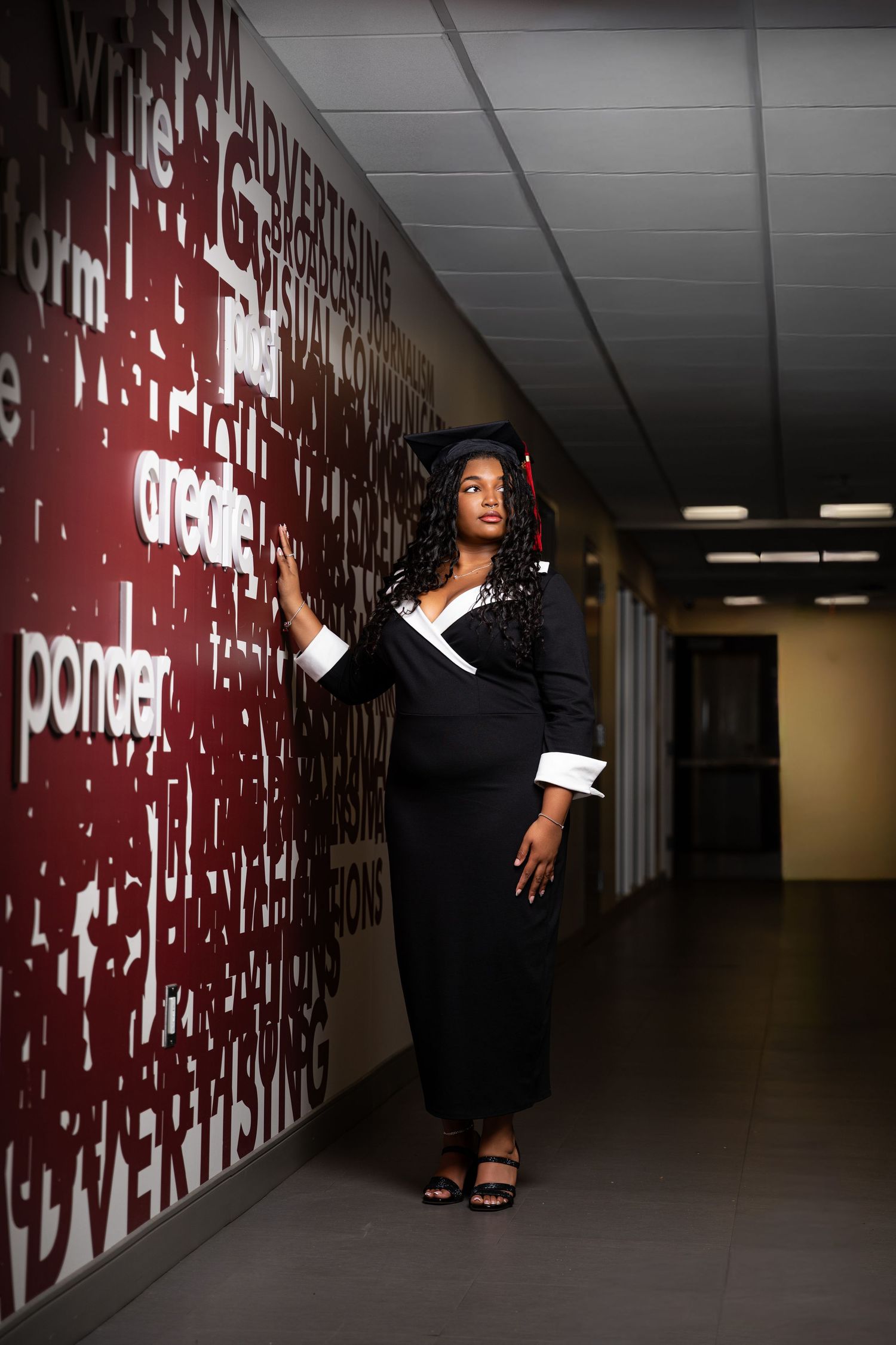 Graduate in black cap and gown posing in a hallway with a red word-art wall featuring advertising terms.