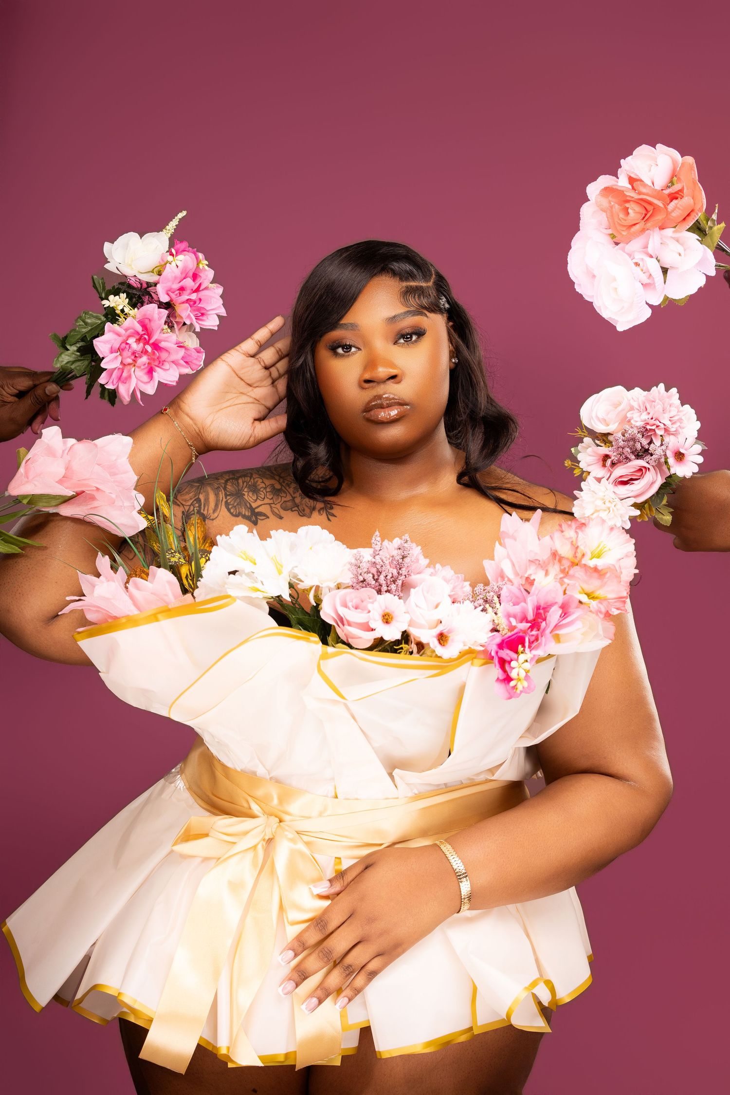 Plus-size woman in white and gold dress surrounded by pink flowers against a mauve background.