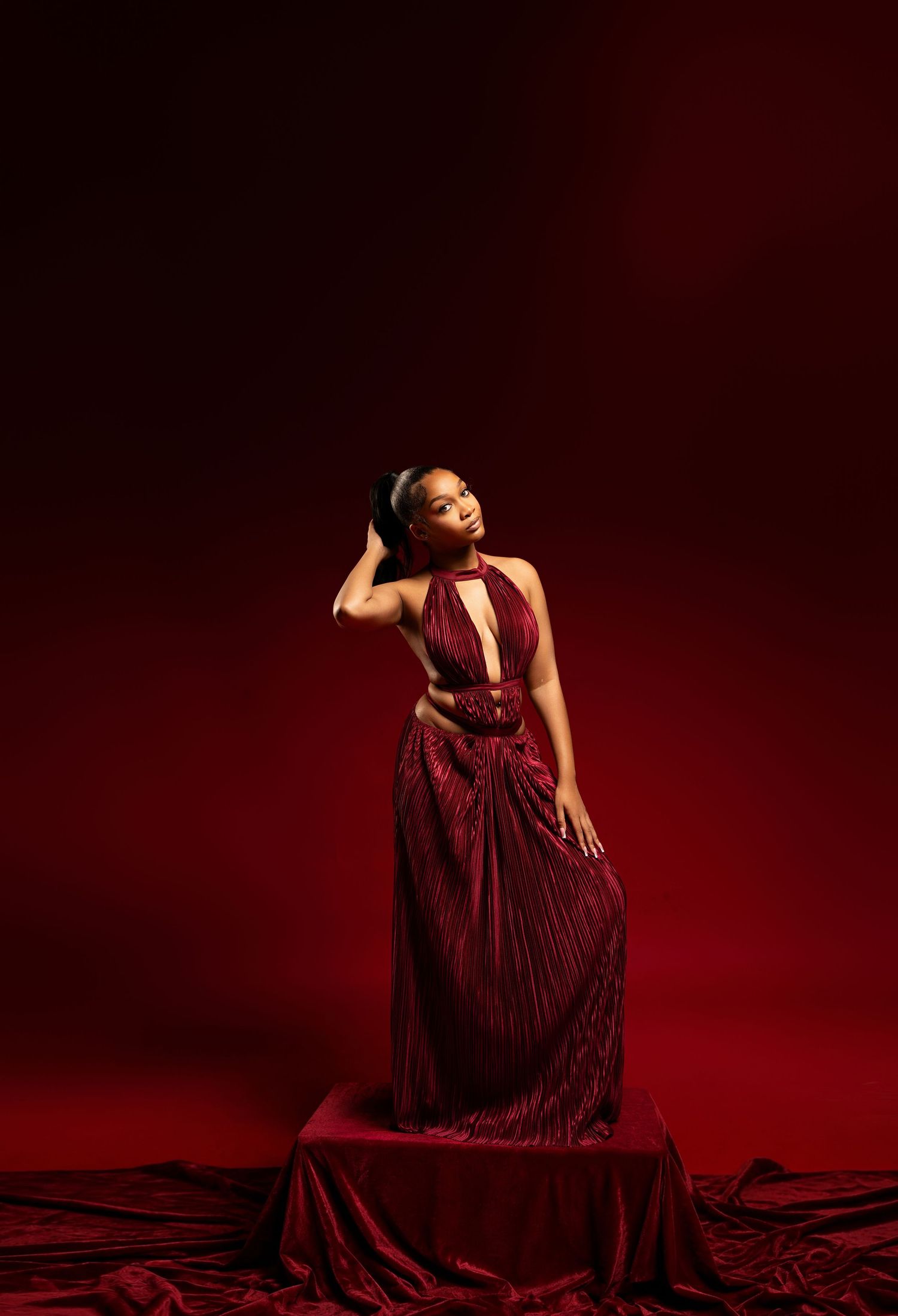 A woman in a stunning deep red halter gown poses elegantly against a dramatic crimson studio background.