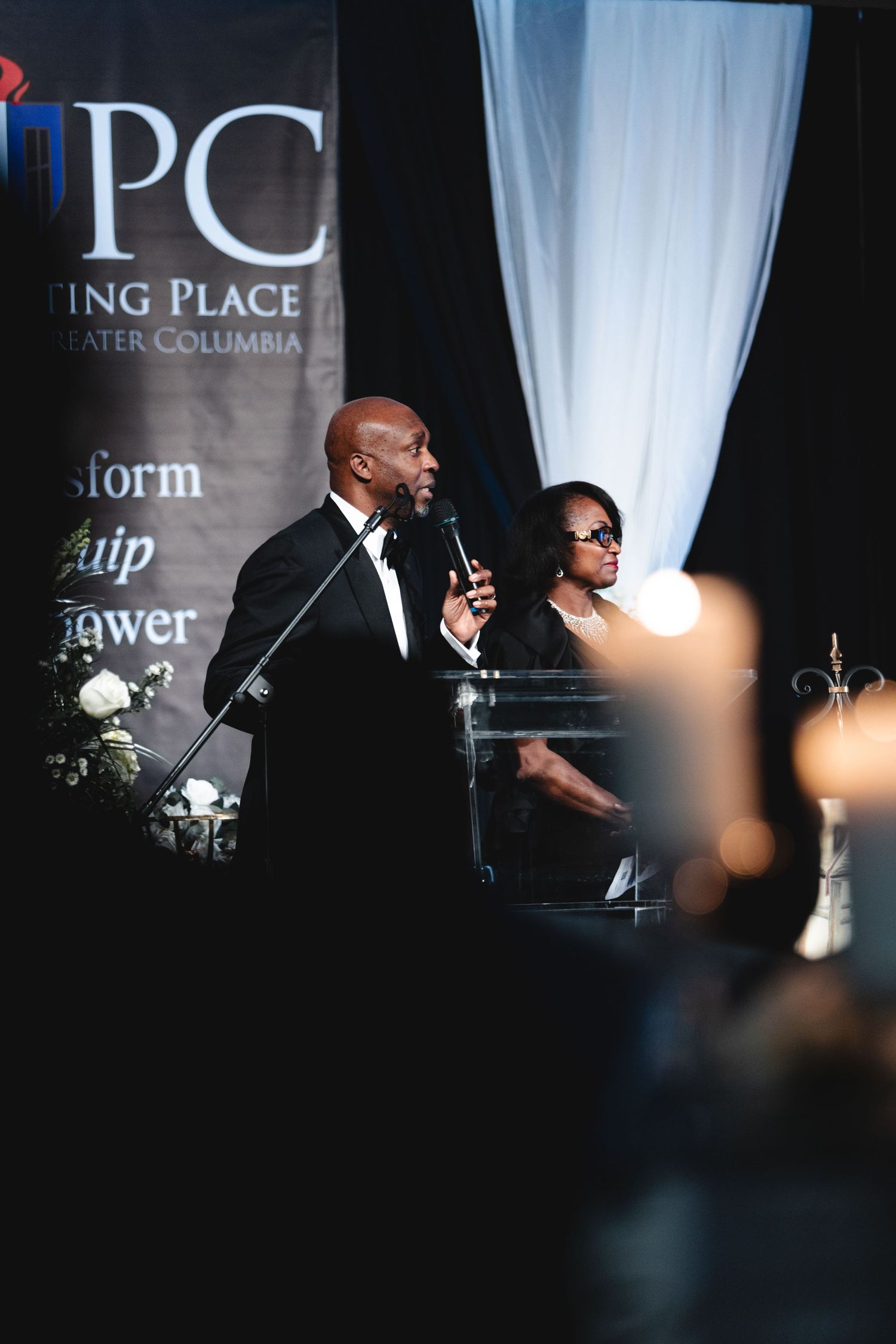 A performer speaks on stage at a formal event with dramatic lighting and white curtain backdrop.