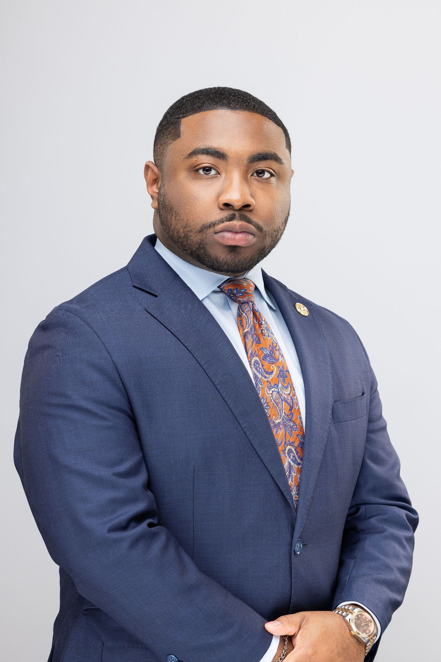 Professional headshot of person in navy suit and orange patterned tie against light background.