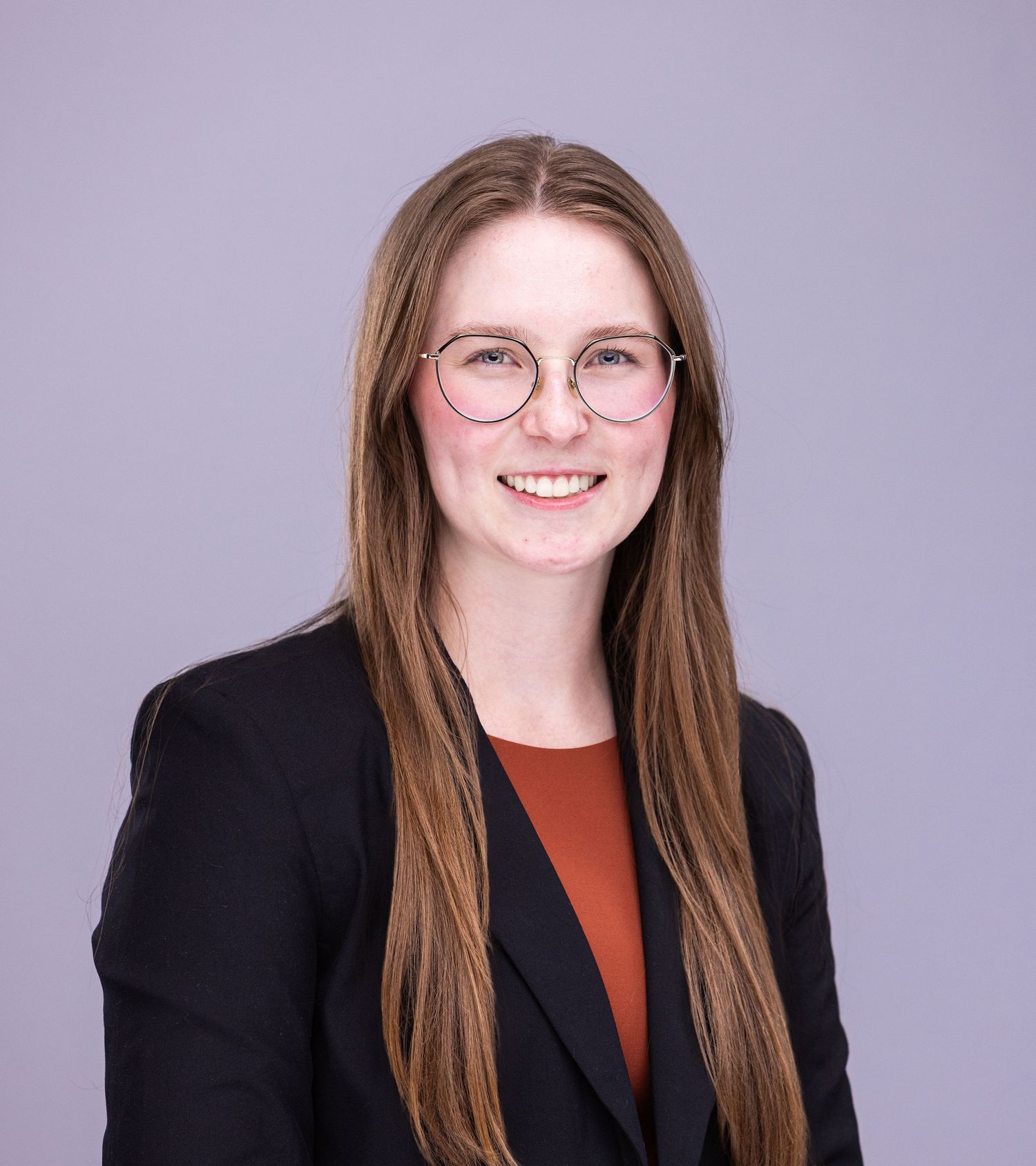 Professional headshot of person wearing glasses, black blazer and rust-colored top against purple background.
