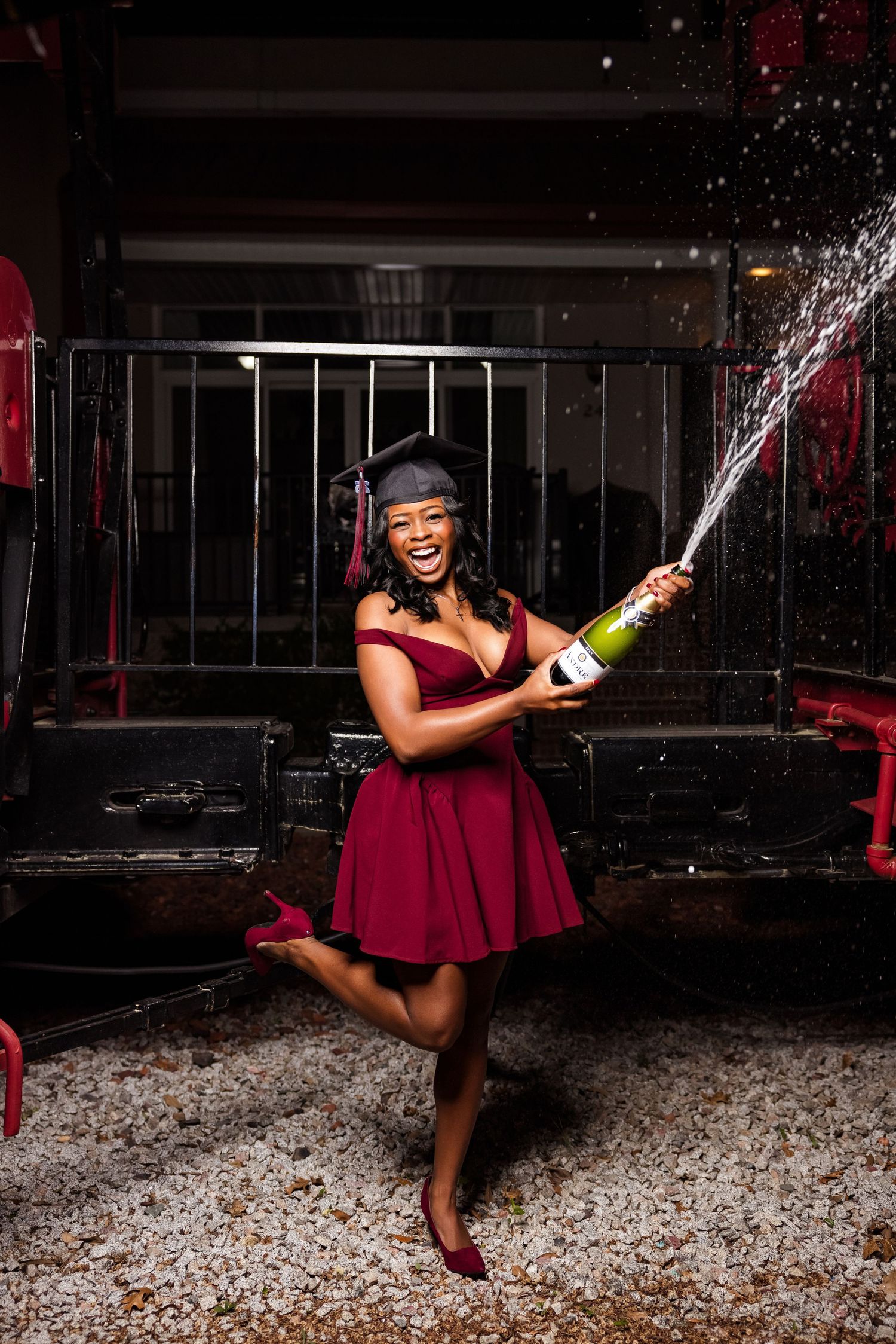 Graduate in red dress sprays champagne while celebrating in festive indoor setting.